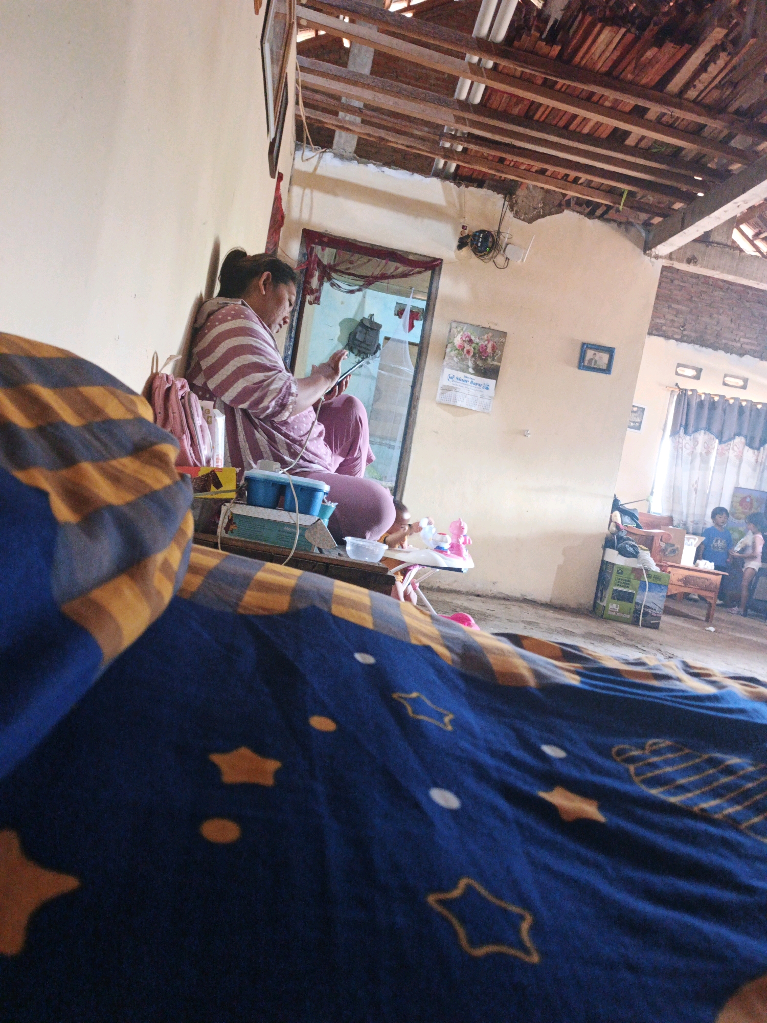 The image depicts an indoor scene in what appears to be a residential space in Bumiharjo, Indonesia. In the foreground, a blue and yellow striped blanket covers a surface, partially obscuring the view. To the left, a woman with her hair pulled back in a ponytail is seated against a cream-colored wall. She is wearing a striped pink and white shirt and pink pants, and is looking at a smartphone in her hands. A pink backpack is leaning against the wall near her. In front of her are various items on a surface, including blue and green boxes, and some small containers.
The background reveals an unfinished ceiling with exposed wooden beams and roofing materials. A doorway with a red curtain leads into another area. On the wall near the doorway, a framed picture of flowers and a calendar are visible. Further into the room, a wooden table and chairs are present, with two children standing near the table. One child, wearing a blue shirt, looks towards the camera, while the other child, wearing a white shirt, is partially visible. A large cardboard box is on the floor to the left of the table. The overall environment suggests a humble, possibly rural, dwelling with signs of ongoing construction or disrepair in the ceiling area.