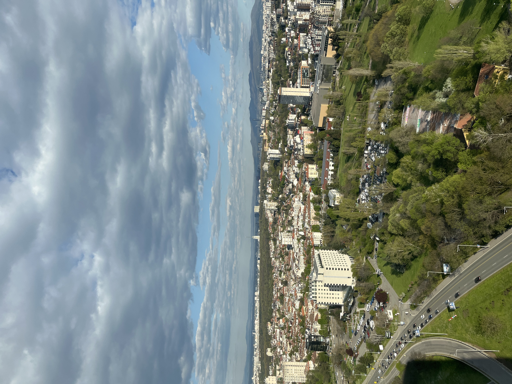 This aerial view captures a wide urban panorama of Sofia, Bulgaria, looking towards the Vitosha mountain range in the distance. The image, originally rotated 90 degrees clockwise, presents a cityscape framed by nature.
The upper portion of the scene is dominated by a dramatic, partially cloudy sky. Large, dense grey clouds with visible textures stretch across, interspersed with clear patches of blue sky, suggesting dynamic weather conditions during daytime.
Below the sky, a prominent mountain range forms the distant horizon line, partially veiled by a light atmospheric haze. At the base of these mountains, and extending across the middle ground, is the sprawling city. It features a dense collection of buildings, including many residential structures with red-tiled roofs, characteristic of European cities. These are interspersed with taller, more modern white or light-colored multi-story buildings, likely commercial or larger apartment complexes.
The foreground is characterized by extensive green spaces. A large, lush park or forested area with numerous trees, showing various shades of green, borders the urban development. Within this park, a concrete amphitheater-like structure is visible, adorned with colorful, abstract graffiti. Several roads traverse the scene; a multi-lane thoroughfare with a central median is busy with vehicular traffic, and a distinctly curved road section is prominent. Cars are seen both moving and parked along these routes and in adjacent parking areas. A large, modern white building with a gridded facade stands near one of the main roads.
A significant location cue is visible on a billboard-like structure next to the main road, where the word "SOFIA" is clearly legible in large white capital letters on a dark background. The overall impression is that of a bustling city integrated with substantial natural green areas, set against a mountainous backdrop.