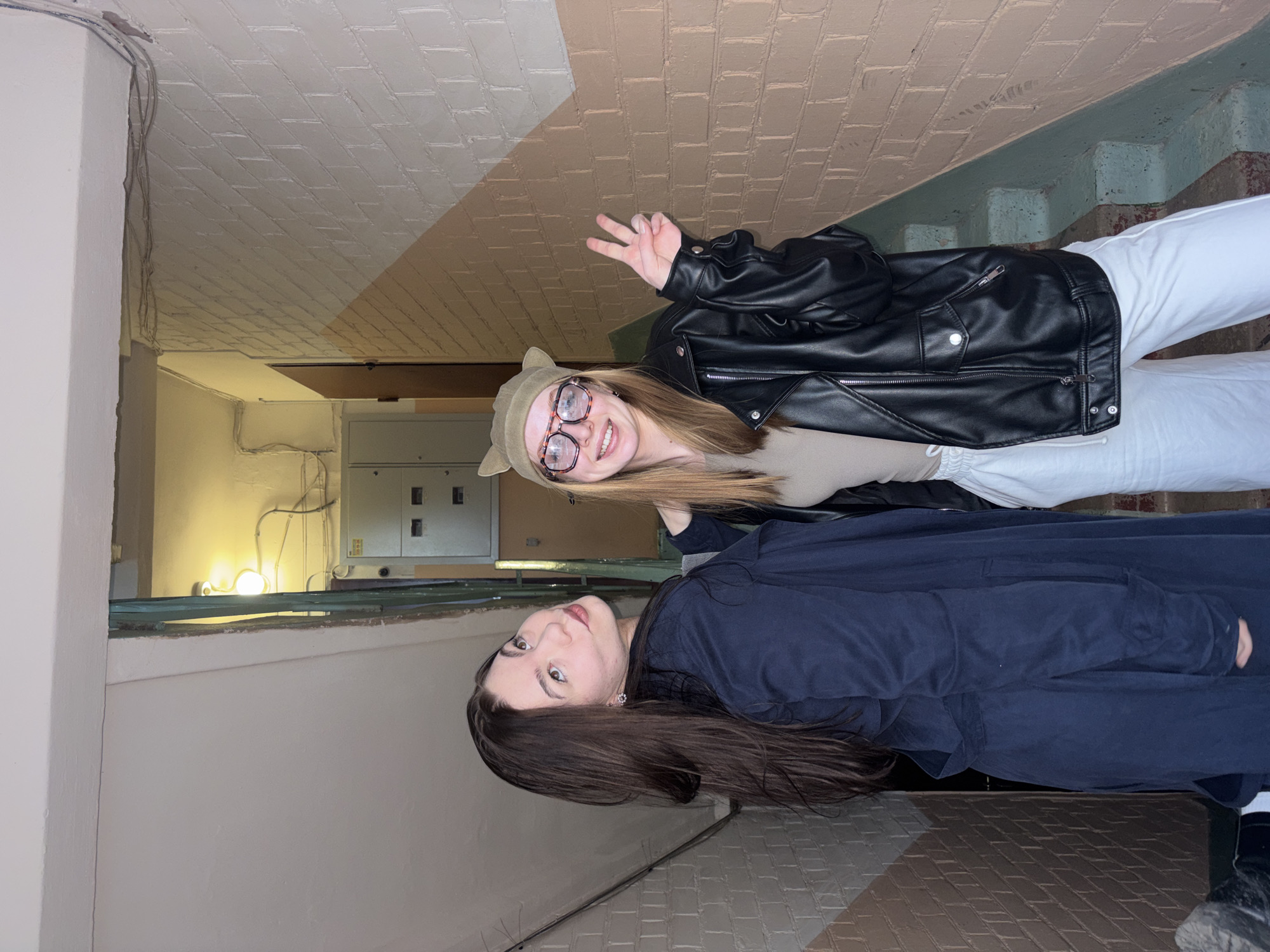 Two young women are posing in what appears to be the stairwell or entrance of an apartment building in Perm, Russia. One woman wears a black leather jacket, white pants, and a hat with cat ears. She is holding up a peace sign. The other woman is wearing a dark blue coat. The stairwell has tiled walls and ceilings. Wires run along the ceiling. A staircase is visible behind the woman in the leather jacket.
