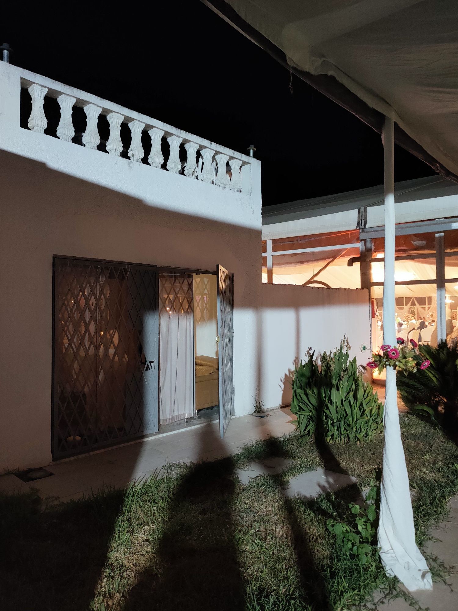 This image depicts the exterior of a building at night, situated in the city of Mornag, Tunisia. The scene is illuminated by artificial light, creating strong shadows. In the foreground, a patch of dry grass and dirt is visible, casting long shadows. To the right, a white cloth is draped over a pole, adorned with a small cluster of pink flowers. Further back, a section of the building features a white, textured wall with a decorative balustrade running along its top edge. Below the balustrade are two sliding doors, partially open, secured by metal security grates. Through the grates and open doors, the interior of a brightly lit room can be seen, featuring beige curtains and what appears to be a large sofa or seating area. Behind this initial structure, a larger building with a translucent roof and extensive glass windows is illuminated from within, revealing a gathering of people seated at tables, suggesting an event or restaurant. Lush green plants are situated in front of the glass structure. The sky above is dark and appears clear.