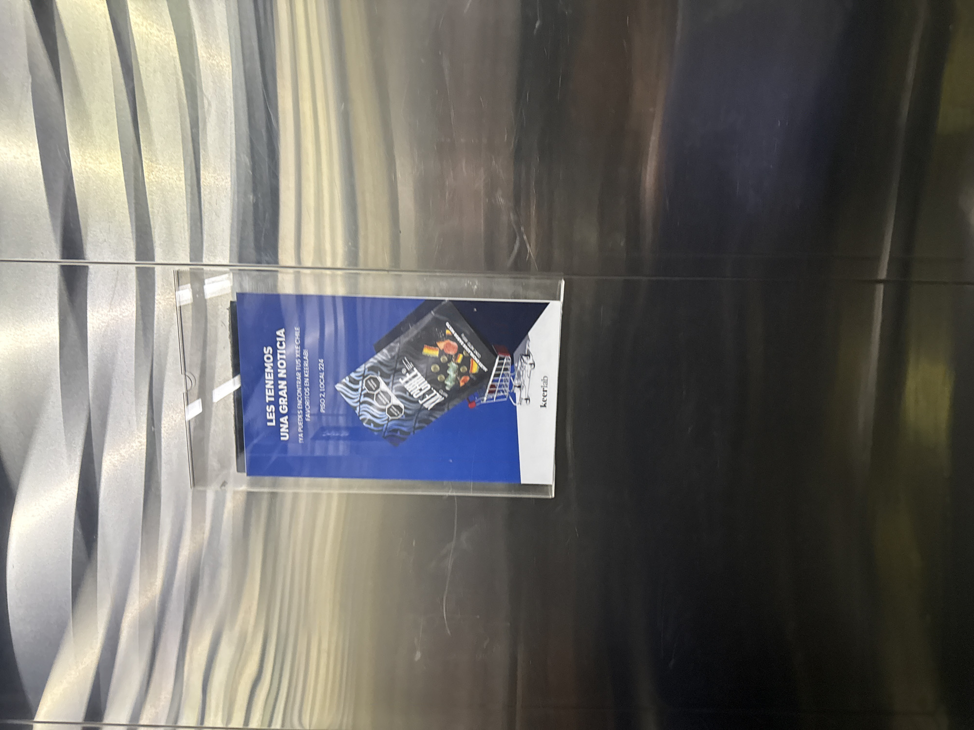 The image shows a promotional flyer attached to a metallic surface, possibly a wall or door. The flyer advertises "Xile Chile" products at Keerlab, located on the 2nd floor, Local 224. It is likely in San Bernardino Tlaxcalancingo, Mexico, given the Spanish text on the flyer and the location mentioned on the flyer. The metallic surface is reflective with a patterned texture in some areas.