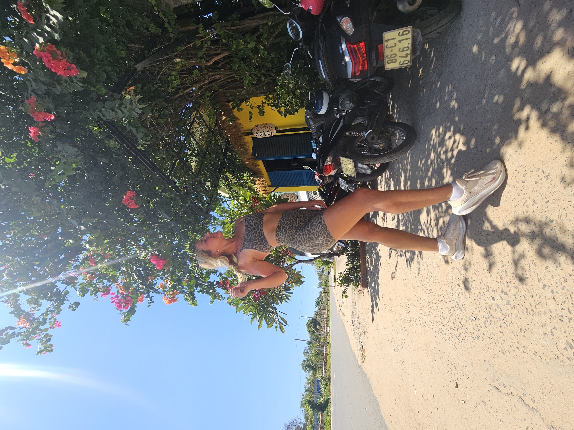 In Mũi Né, Vietnam, a woman with blonde hair, wearing a leopard-print sports bra and shorts, stands on a sandy roadside under a tree with pink and orange flowers. She is looking to her right, with her right arm raised and hand touching her hair. To her right, parked motorcycles are visible, one with a yellow license plate displaying "86-C1 646.16". A yellow building with blue shutters is behind the motorcycles. The background shows a paved road, palm trees, and a clear blue sky with sunlight streaming from the left.