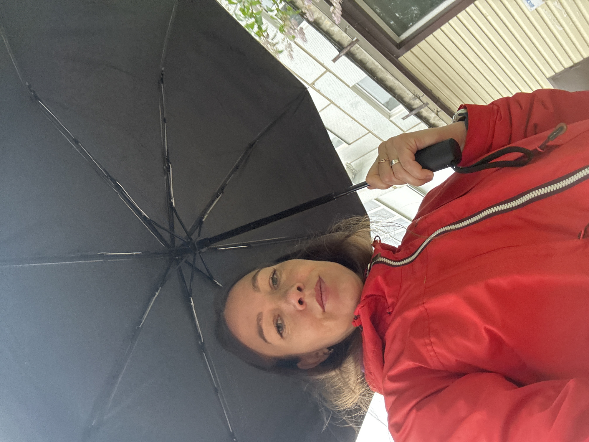 A woman stands beneath an open black umbrella. She wears a red jacket with a white-and-black stripe. She holds the umbrella's black handle in her hand, which is adorned with two rings. Above her, a building with light-colored siding and a window can be seen. The setting is likely an urban area based on the architectural details, but a precise location within Chaikovsky, Russia, cannot be determined from the image alone.
