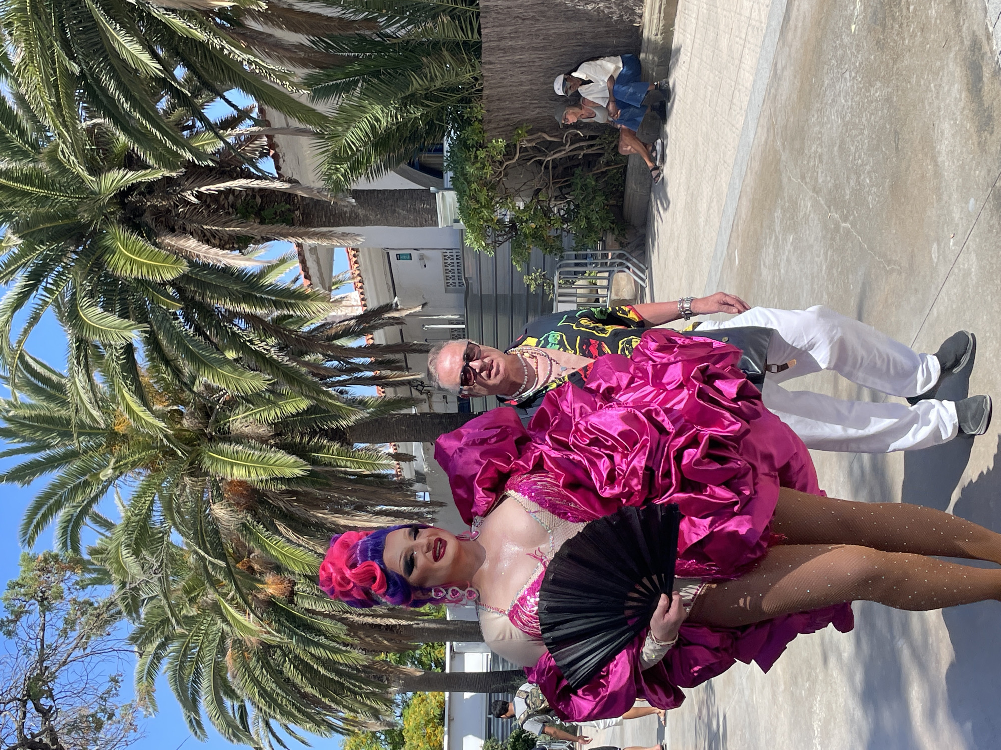 A drag queen in a vibrant pink ruffled costume and purple wig poses with a black fan in front of palm trees and a white building in Sitges, Spain. She wears sparkling stockings and dramatic makeup. Beside her stands a man in a colorful shirt, sunglasses, and multiple necklaces. In the background, two individuals are seated on a ledge near a wooden fence. The scene appears to be outdoors, likely during a sunny day.