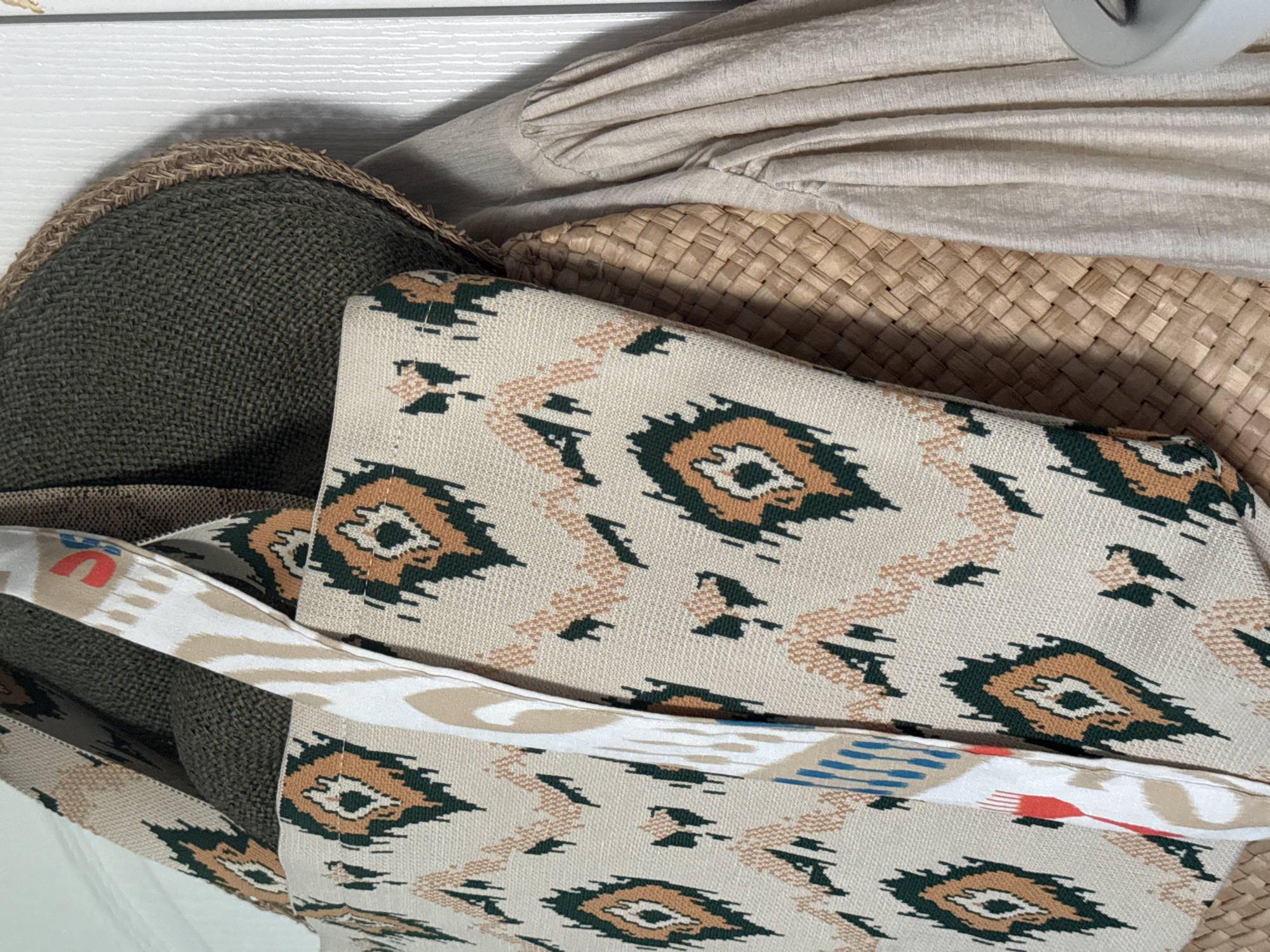 The image shows a close-up of several objects in a seemingly random arrangement. There's a dark green woven hat with a rough edge, a tan woven bag or basket, a folded light beige garment, and a beige patterned fabric bag with a wide strap featuring colorful accents (red and blue). The scene appears to be indoors, possibly in a storage area. There is no information to suggest the location is in the city of Kazan, Russia.