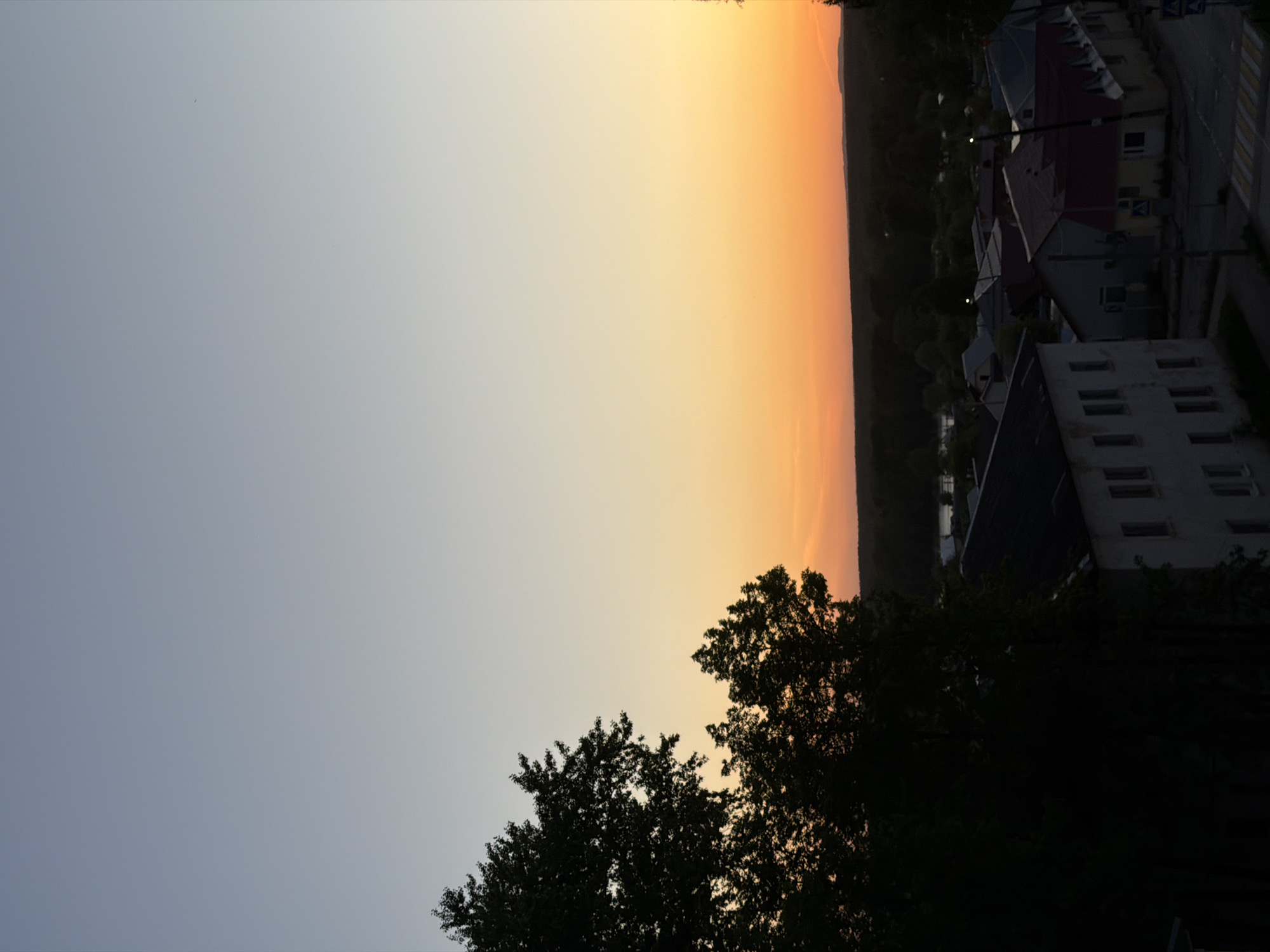 The image depicts a twilight or sunset scene in Chaikovsky, Russia. The sky transitions from a light blue-gray on the left to an orange-yellow towards the horizon in the center. Silhouetted treetops occupy the lower right of the image. In the right side, buildings are visible, including a white apartment building with multiple windows. The image is turned to the side, with the horizon going along the length of the photo instead of the width.