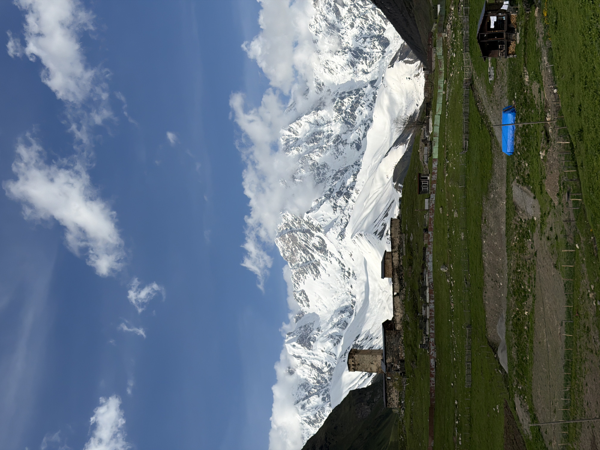 The image displays a vertical panorama of a mountainous landscape in Ushguli, Georgia. The scene is divided into two main sections: the sky and the mountains, and the village and surrounding terrain.
The upper left portion of the image is dominated by a vibrant blue sky with scattered white clouds. The clouds are wispy and spread across the sky, indicating a bright and likely sunny day.
The right side of the image features a towering, snow-capped mountain range. The snow covers the peaks and upper slopes, creating a stark contrast with the darker rock formations visible in places. Some clouds are clinging to the mountains, suggesting potential weather patterns or the altitude of the peaks.
Below the mountains, a green, grassy slope leads down to a small village. Several stone structures, possibly ancient defensive towers or homes, are visible along the hillside. The village appears to be situated in a valley or on the side of a mountain, with agricultural terracing or fenced areas on the grassy slopes. A prominent blue tarp is stretched between poles in the village area, possibly for shelter or protection. A dark wooden building with a balcony and stacked firewood is also visible, suggesting a dwelling or small guesthouse. Wooden fences and pathways mark the landscape.
The image conveys a sense of rugged natural beauty and remote habitation. The time of day appears to be daytime, with ample sunlight illuminating the scene. The weather seems clear, though the presence of clouds around the mountain peaks might suggest cooler temperatures at higher elevations. The location cues, such as the distinct stone towers and the overall mountainous terrain, point to a location like Ushguli, known for its medieval architecture and dramatic Caucasus mountain setting. No visible text is present in the image.