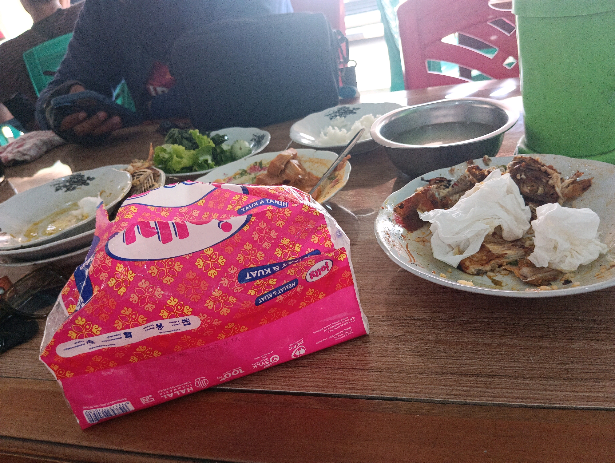 The image shows a partially eaten meal on a wooden table. In the foreground, a pink and white patterned tissue packet is prominent. Several plates are visible, some containing remnants of food such as rice, vegetables, and what appears to be fried chicken or fish. A metal bowl of liquid, possibly water or soup, is also present. In the background, a person wearing a dark blue shirt is partially visible, seated at the table and holding a smartphone. The environment suggests an indoor setting, possibly a restaurant or home, with a glimpse of red chairs and a green container in the background. The context provided indicates this scene is located in Belitang, Indonesia.