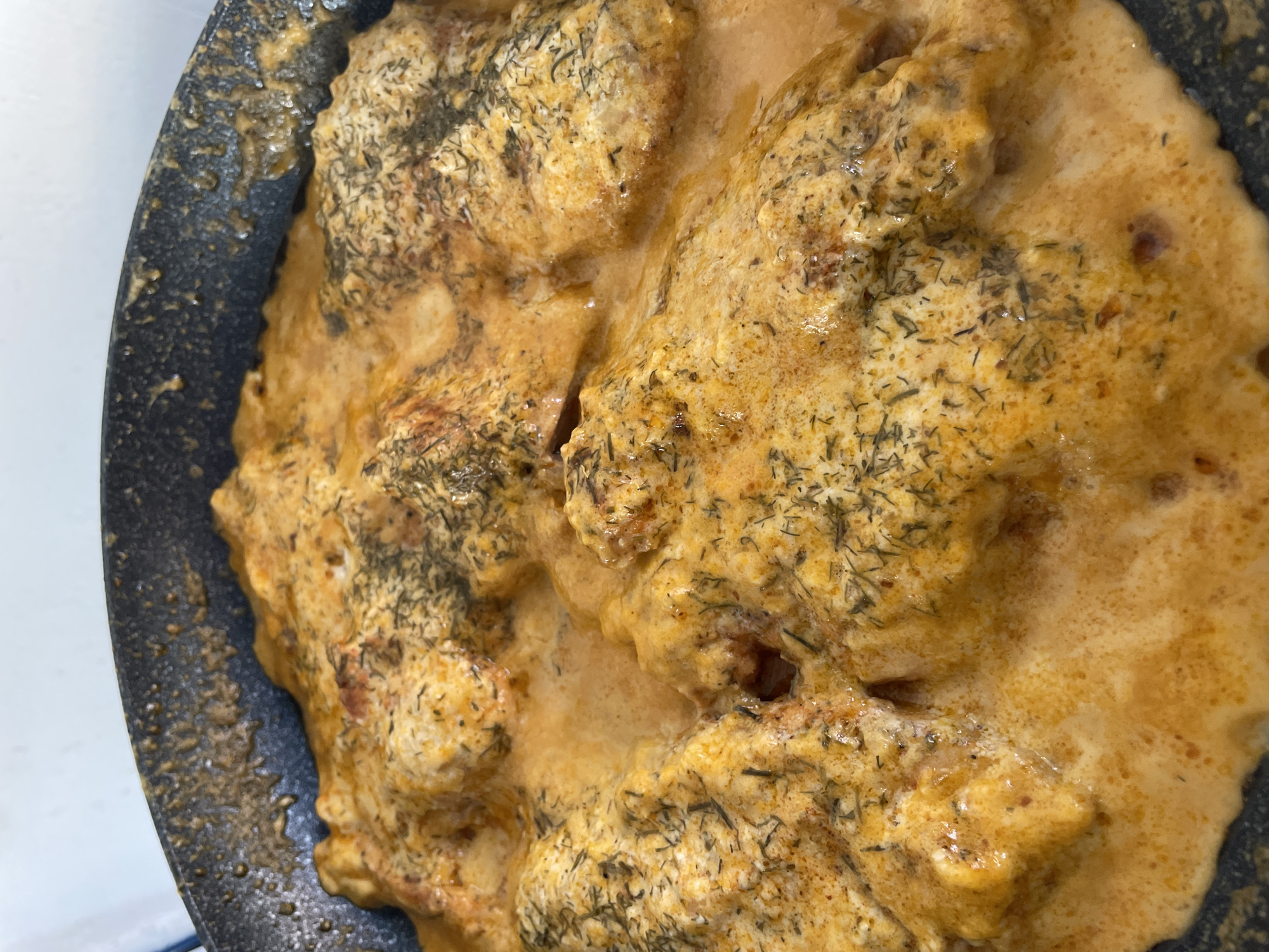 A close-up, overhead shot shows pieces of meat, possibly chicken, simmering in a creamy, orange sauce in a black frying pan. Dried herbs, likely dill, are sprinkled over the meat and sauce. The pan is dark and appears to be seasoned, with some food residue visible on its inner rim. The lighting is bright, creating a warm, inviting glow over the dish. The setting is likely a kitchen in Skala Avdiron, Greece, though no specific landmarks or environmental details confirm this location.