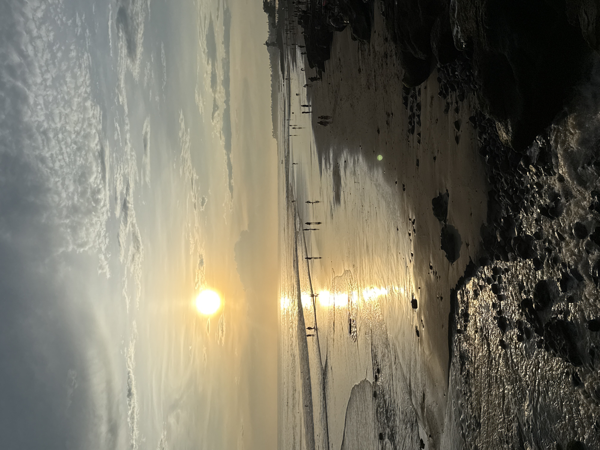 The image depicts a picturesque sunset scene at a sandy beach, likely in North Kuta, Indonesia. A brilliant, golden sun hangs low on the left side of the frame, casting an intense, warm light across the entire landscape. The sky above is a blend of soft grey and white clouds, with clearer, golden-tinged patches near the horizon and surrounding the sun.
The wet sand of the beach strongly reflects the sunlight, creating a luminous, golden path stretching towards the viewer and leading to the horizon. Numerous silhouetted figures of people are scattered across the expansive beach, walking along the water's edge and on the wet sand. Their forms are dark against the bright reflections, indicating the low angle of the sun. Some individuals walk alone, while others appear in small groups.
On the right side of the image, dark, rugged rock formations or cliffs rise prominently, appearing almost black due to the backlight, creating a stark contrast with the bright sky and water. These rocks show textured surfaces, some glistening, suggesting they are wet from the tide or spray. The ocean meets the shore in gentle, reflective waves. The overall atmosphere is one of serene natural beauty during the golden hour, with no visible text.