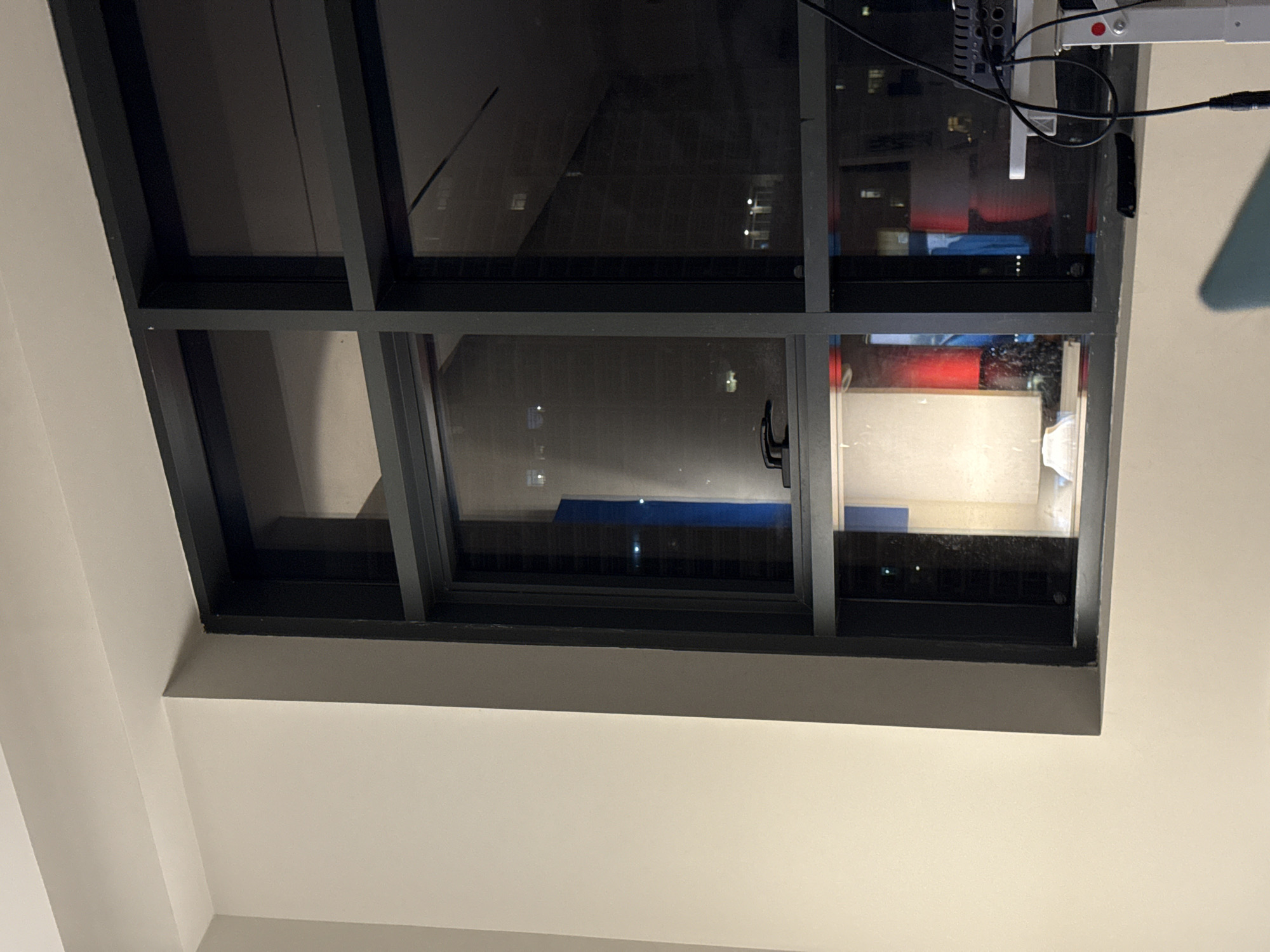 The image shows a ceiling with a large window that appears to be a skylight. The window is divided into several panes by dark, rectangular frames. Through the window, one can see the night sky, with reflections of lights from buildings. Some of the panes show the interior of a room, with shelves and electronic equipment visible, including what appears to be an audio mixer. There are also some cables connected to the equipment, and a white light fixture with a red button is attached to the ceiling above the window. The overall scene is dimly lit, suggesting it is nighttime. The location is identified as Dubai, United Arab Emirates, which implies an urban setting with modern architecture. There is no specific activity or event clearly depicted, but the presence of the equipment and cables might suggest a setup for entertainment or production. The mood is somewhat muted due to the low light conditions. No discernible text is visible in the image.