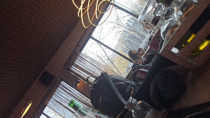 The image captures the interior of what appears to be a cafe or restaurant in Varna, Bulgaria, seen from a slightly elevated, diagonal perspective. The precise location within Varna is not visually discernible.
In the immediate foreground, a clear glass bottle with a cork stopper is visible, alongside a segment of a wooden crate or holder containing other bottles, one of which contains a yellow liquid. A dark, out-of-focus object, potentially part of the viewer's clothing or another item on the table, occupies the bottom portion of the frame.
Mid-ground left displays a partially visible green glass bottle. A black backpack with zippers rests on a seat or bench to the right of the green bottle. Further into the mid-ground, a female individual with long, dark, curly hair is seated at a table near a large window, oriented away from the camera. She is dressed in a light-colored top and is engaged with a small object held in her hands, likely a mobile phone. Her seating area is adorned with patterned cushions. Above this area, a prominent, modern light fixture composed of multiple glowing concentric rings illuminates the space.
The background is dominated by large windows that offer a view of an outdoor environment. Bare tree branches are visible, suggesting a colder season. Beyond the trees, a distinctive architectural feature of numerous vertical, light-colored slats or pipes is present. Distant hills or landscape features are visible under a bright sky.
The indoor setting includes a dark wooden paneled ceiling, sections of exposed brick walls, and sheer, light-colored curtains draped over the windows. Artificial lighting fixtures contribute to the interior illumination.