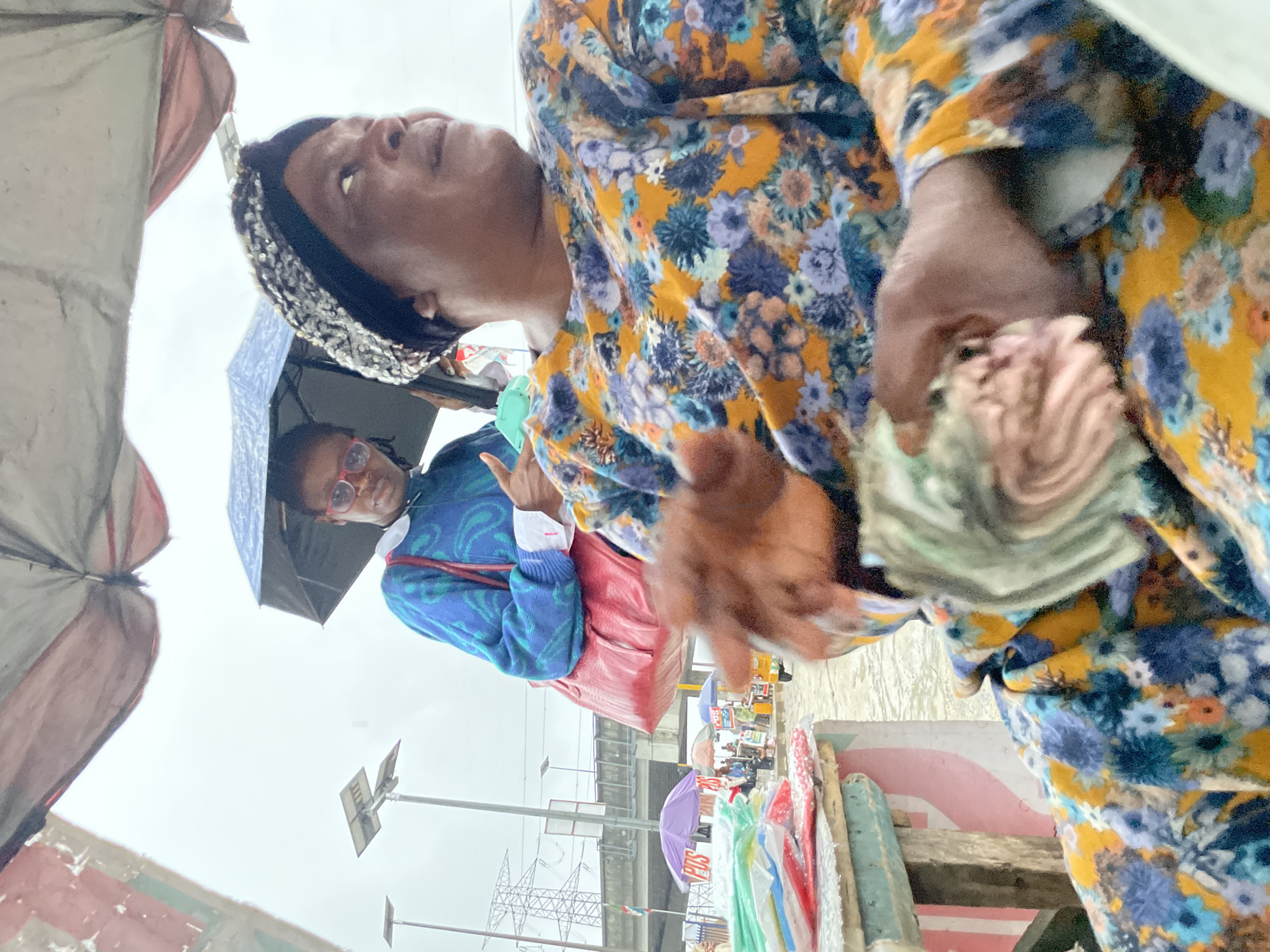 The image depicts an outdoor scene in Lagos, Nigeria, likely a marketplace given the presence of a vendor and various goods. The foreground is dominated by an adult woman, wearing a floral-patterned dress and a sparkly headwrap. She is holding a fan of banknotes in her hands, suggesting a transaction or counting money. To her left, a table is laden with colorful plastic bags, possibly containing merchandise.
In the middle ground, a younger woman, wearing a blue sweater with a white collar, glasses, and a red shoulder bag, holds a black umbrella, indicating potentially rainy weather. She appears to be looking towards the camera and giving a thumbs-up gesture. Behind her, other market stalls with umbrellas and some people can be seen, along with a yellow tricycle.
The background reveals more of the urban environment of Lagos, including elevated structures like a bridge or highway, power pylons, and streetlights. The overall atmosphere suggests a busy, everyday scene in the city.
