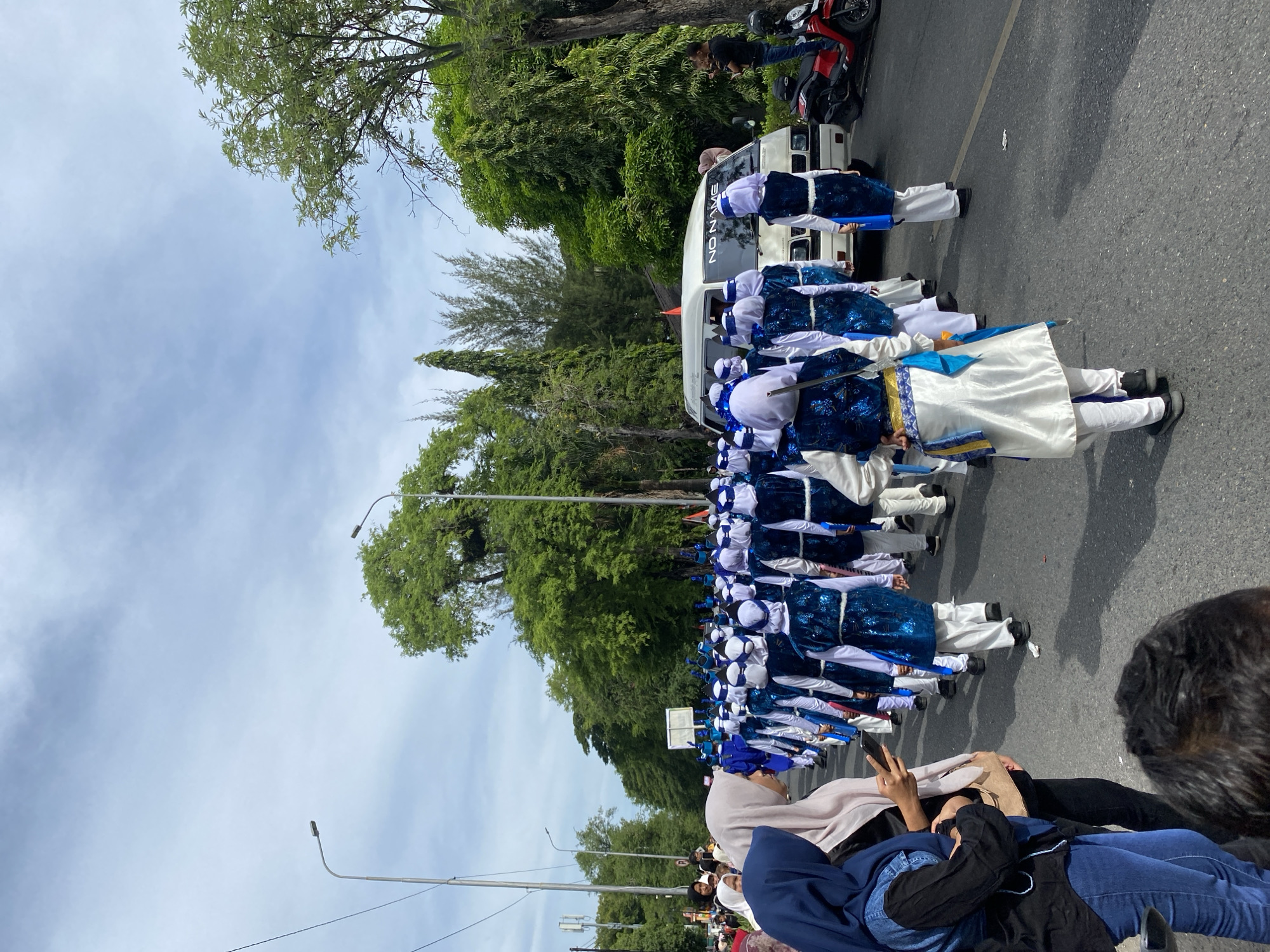 The image depicts a street scene in Banda Aceh, Indonesia, on a cloudy day. A marching band, composed of individuals in matching blue and white uniforms with ornate blue vests, is proceeding down a paved road lined with lush green trees. The band members are wearing white pants and some are seen holding blue instruments or flags. In the foreground, several spectators are visible. On the left, a woman wearing a blue hijab and a black shirt with denim jeans is looking at her phone, with another woman in a light-colored hijab next to her. Further down the street, a white van with "NO NAME" written on its windshield is part of the procession. A man in a black shirt and jeans is standing on a red motorcycle to the right of the van, possibly playing a violin. The overall atmosphere suggests a public event or parade.