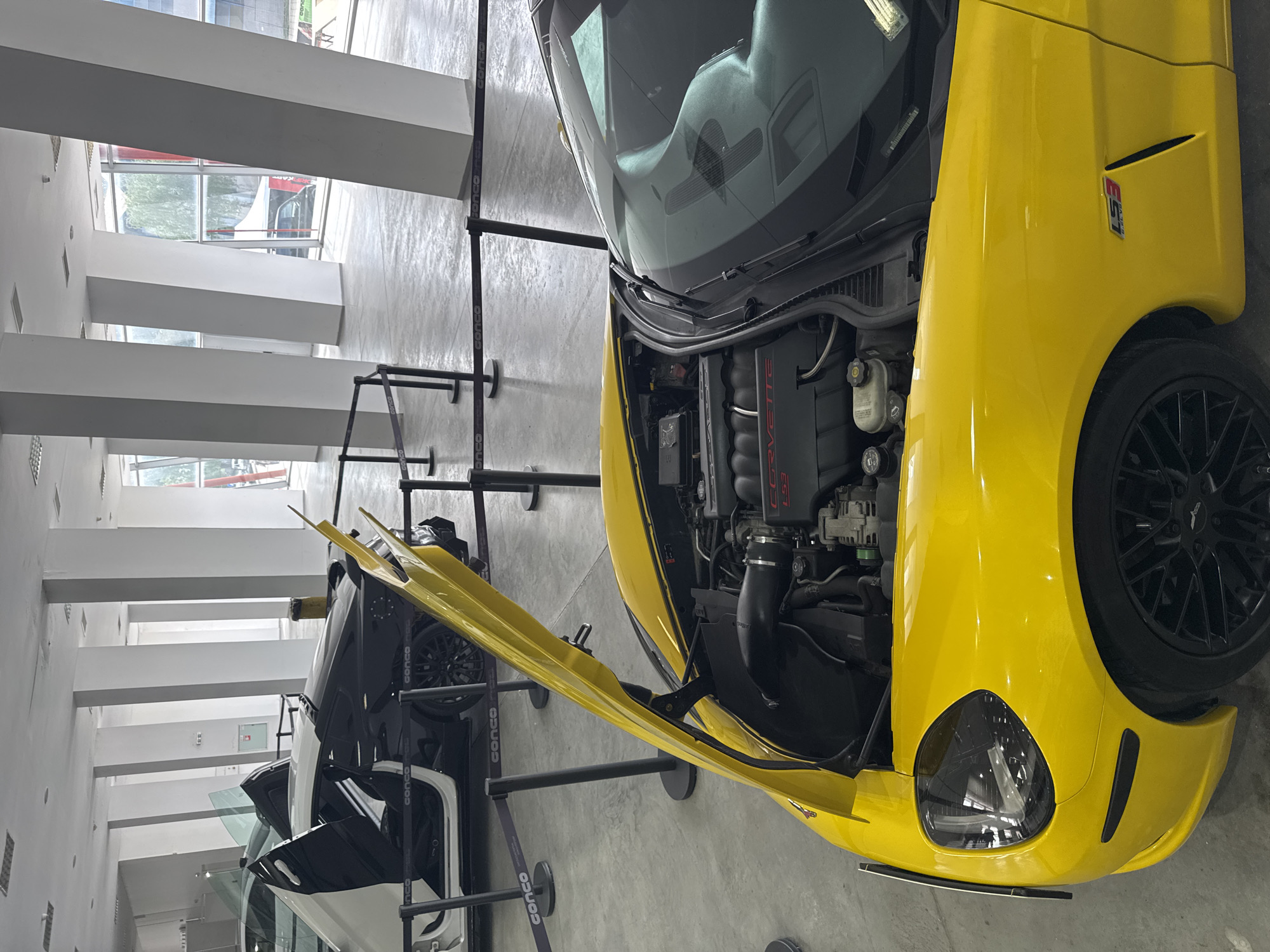 The image shows a yellow Chevrolet Corvette with its hood open, parked indoors. The car's engine is visible. In the background, there are other cars and support structures, suggesting a car exhibition or museum. The location is Plovdiv, Bulgaria, as indicated by the "CONCO" branding on the support structures, which is a company known to operate in Plovdiv. The environment is a modern building with bright lighting and pillars. No people are visible in the image.