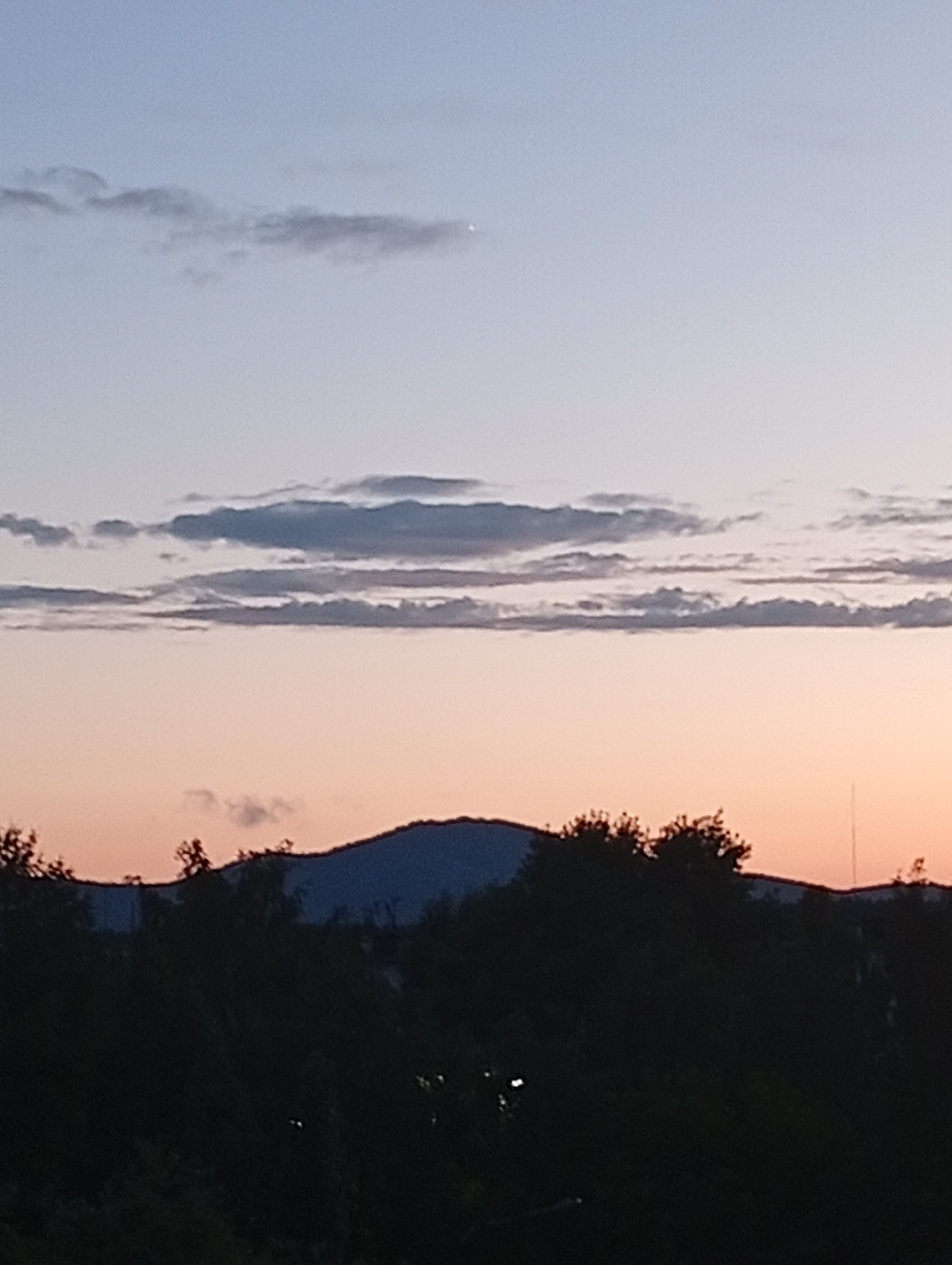 The image captures a twilight scene over Kyshtym, Russia, with a clear horizon dominated by a silhouette of trees and a mountain range. The sky transitions from a soft blue at the top to a warm, peachy-orange hue closer to the horizon, indicating either sunrise or sunset. Scattered across the upper portion of the sky are wispy, dark clouds, adding texture to the vast expanse. A single, bright star or planet is visible in the upper left quadrant of the sky. The foreground is filled with the dark silhouettes of dense foliage and trees, obscuring any finer details of the landscape below. The mountain range in the background is also a dark silhouette against the colorful sky, its peak gently undulating. The overall mood is serene and tranquil, typical of a quiet evening or early morning. There is no visible text in the image.