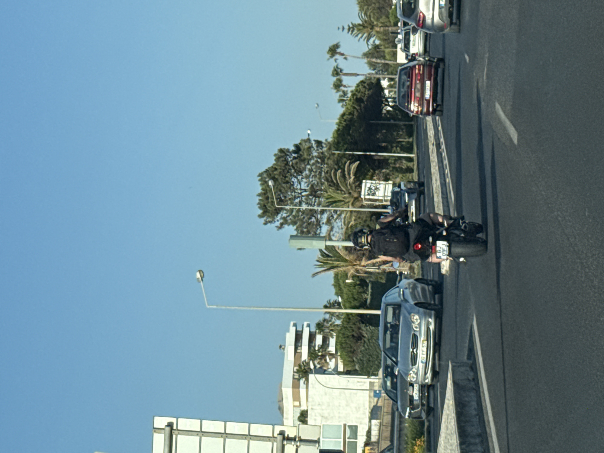 The image depicts a street scene in Oeiras, Portugal, on a clear day with a bright blue sky. In the foreground, a person on a motorcycle, wearing a helmet and a backpack, is riding away from the camera in the right lane of a multi-lane road. The motorcycle's license plate is visible, showing "BJ 01FJ". Several cars are also present on the road. A silver Mercedes-Benz sedan is in the left lane, with license plate "99 CF 57". Behind it, a dark-colored Audi sedan is visible. Further down the road, a red sedan is in the right lane, and a white car is in the left lane.
On the left side of the road, a modern white building with multiple windows is visible, possibly an apartment complex or office building. Further back, there are trees and palm trees, characteristic of the Portuguese coastline. Streetlights are present along the road. The road appears to be a main thoroughfare, with distinct lanes and a concrete barrier separating the lanes. The overall impression is of a typical urban or suburban traffic scene in a sunny climate.