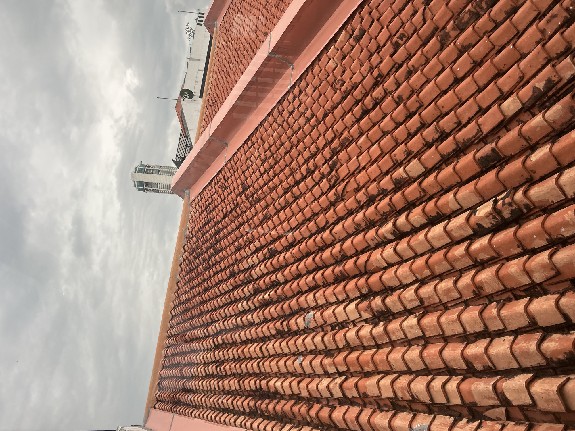 The image captures a view of a red-tiled roof under a cloudy sky, taken from a slightly elevated perspective. The roof is composed of numerous interlocking terracotta tiles, arranged in diagonal rows that recede into the distance. Some tiles show signs of weathering, with darker patches and lighter wear. A pinkish-red metal gutter or flashing runs along the top edge of a section of the roof, following its slope. In the background, other rooftops are visible, along with a tall, modern building with a distinctive green and white top, and another building displaying a circular logo with the letters "CM" inside a green circle. The presence of these distinct buildings, particularly the one with the "CM" logo, strongly suggests the location is within George Town, Penang, Malaysia, likely in an area with a mix of traditional tiled roofs and contemporary architecture. The overall environment appears to be urban, with overcast weather conditions. No people or overt actions are depicted.