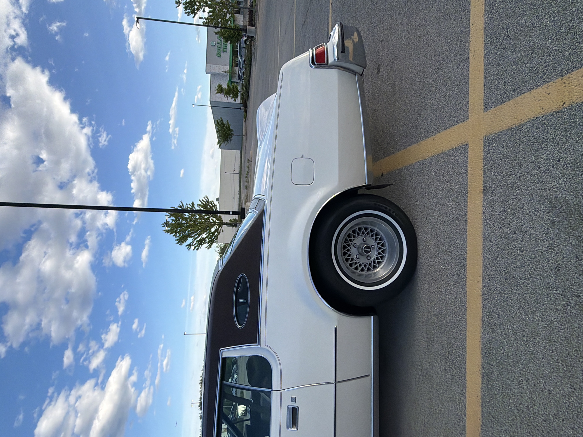 The image shows a white and brown Lincoln Continental parked in a parking lot in Morton Grove, Illinois, United States. The rear portion of the car is visible, showcasing the white body, a brown roof, and a silver wheel. The parking lot is marked with yellow lines. In the background, there is a shopping center featuring a Dollar Tree. The sky is bright with clouds.