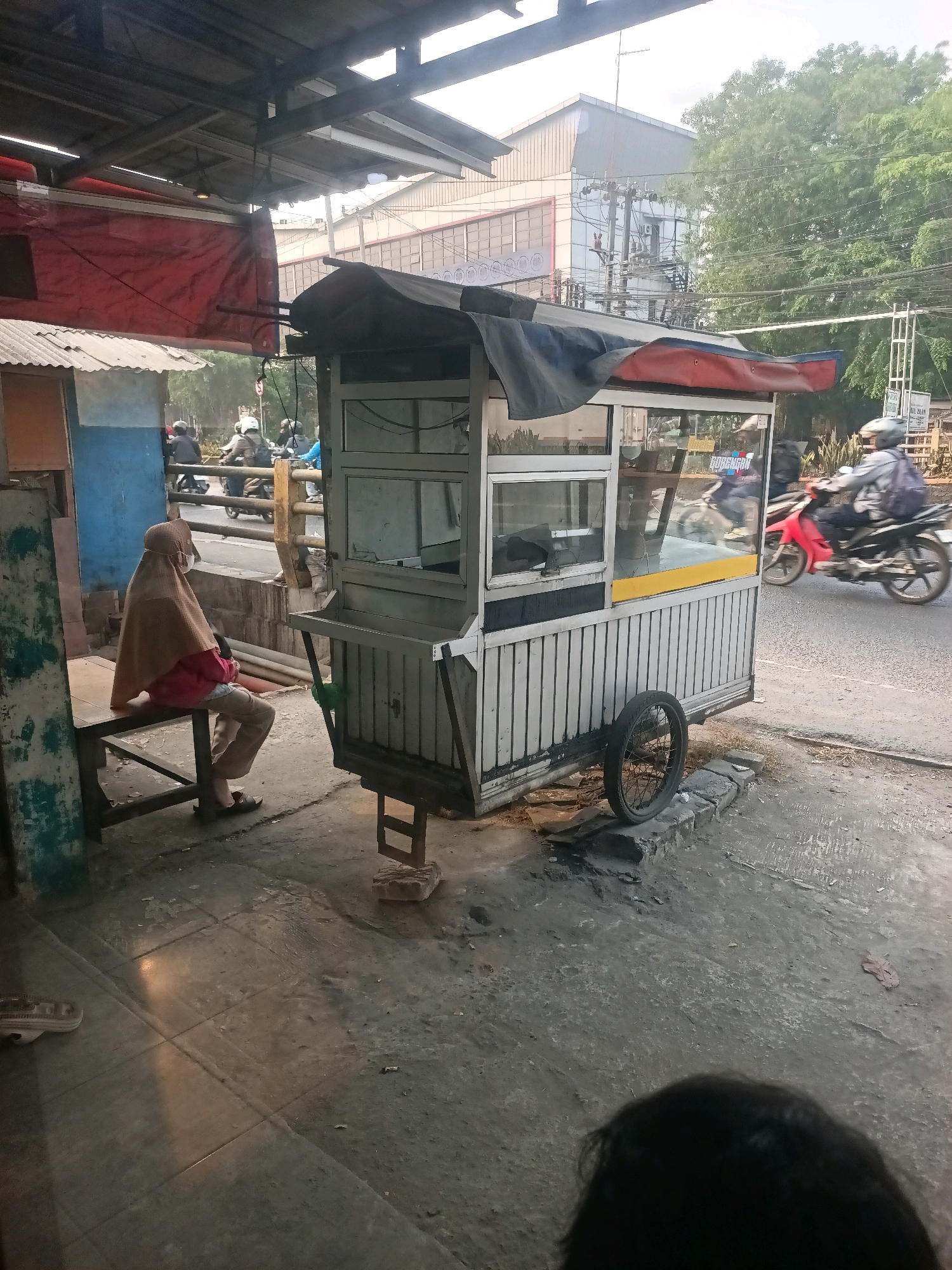 The image depicts a street scene in Gandasari, Indonesia. A food cart with a weathered aluminum frame and a black and red tarp roof is parked on the side of a paved road. The cart has a glass display case on top. To the left of the cart, a person wearing a light brown hijab, a pink shirt, and light-colored pants sits on a wooden bench. They are also wearing a face mask. The background shows a building with a gray facade and trees. Several motorcycles are visible on the road, with one red motorcycle in motion to the right of the food cart. The ground is a mix of concrete and dirt. The overall atmosphere suggests a casual, everyday street setting.