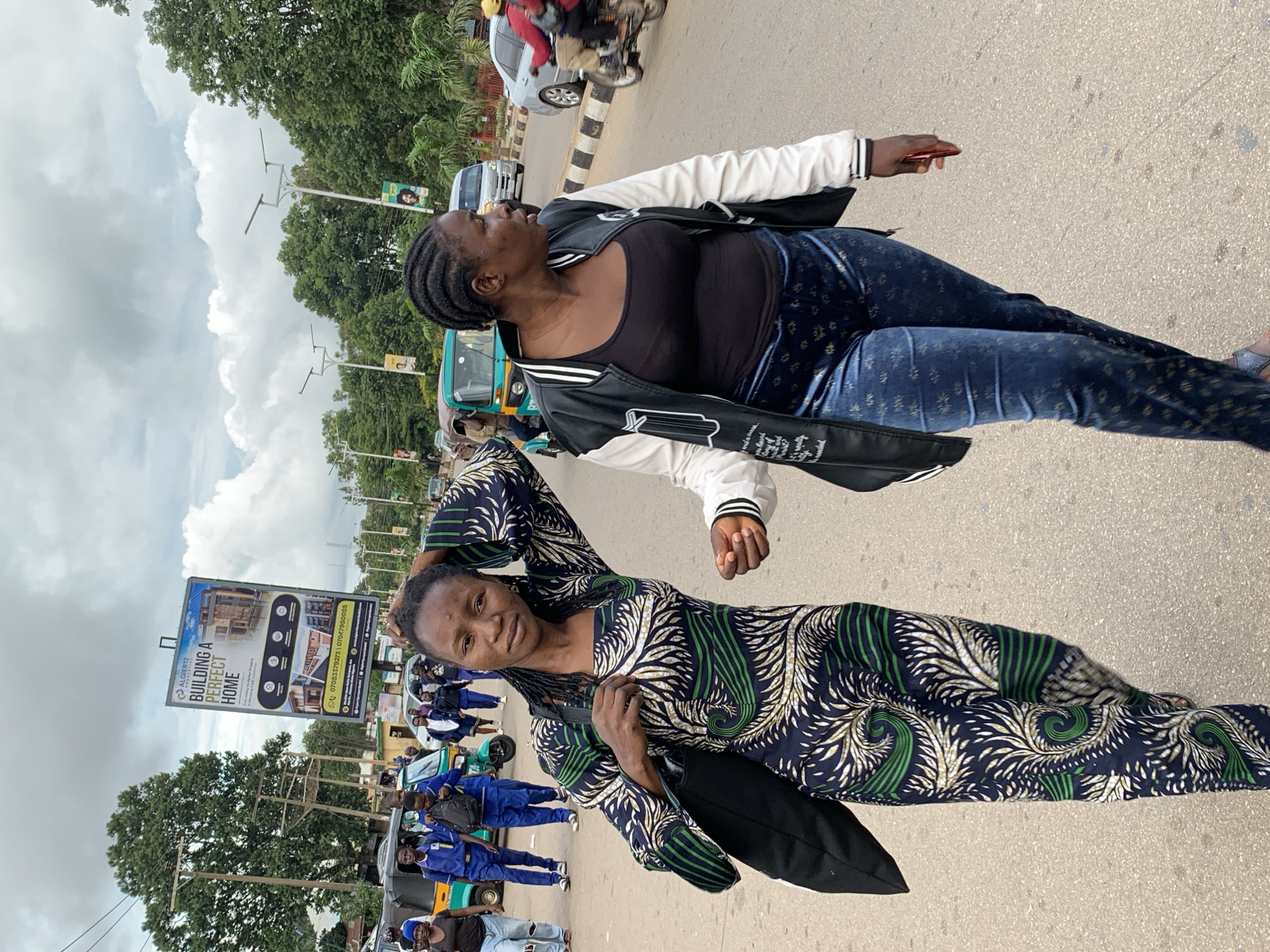 In Barakin Lerawi, Nigeria, two women walk on a paved road under a cloudy sky. The woman on the left, facing the camera, wears a dark blue, green, and white patterned long-sleeved dress and carries a black bag over her left shoulder. Her right arm is raised and her hand is on her head. The woman on the right, facing away from the camera, wears a black tank top, a white and black letterman jacket, and blue patterned pants. She holds a red object in her right hand. Several people and vehicles are visible in the background, including a trishaw, a car, a motorcycle with a rider, and a billboard advertising "ALGIER12 PROPERTIES BUILDING A PERFECT HOME." Trees and utility poles line the sides of the road.