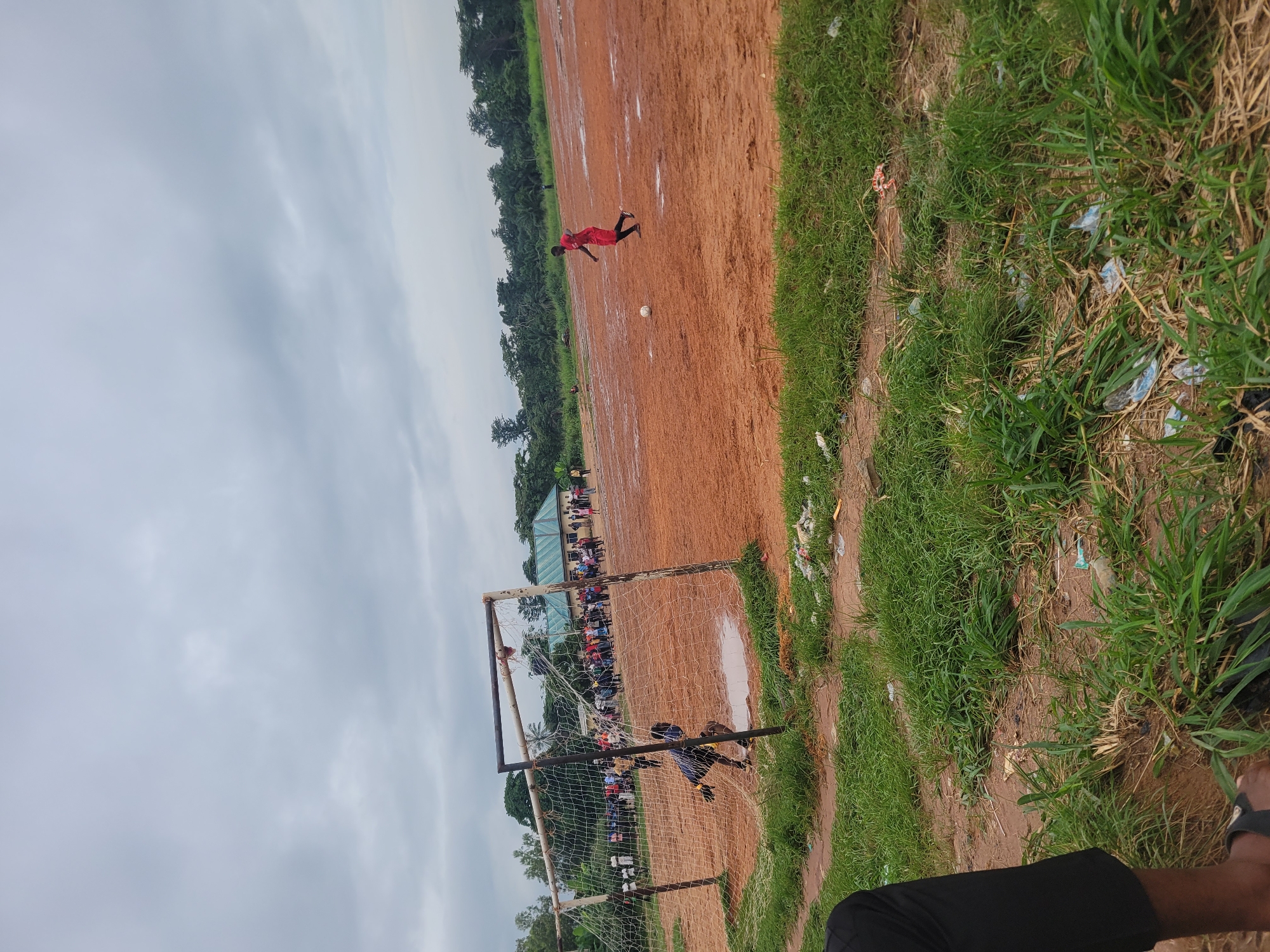 The image depicts an outdoor football match played on a muddy, red dirt field in Egbagu, Nigeria. The sky is overcast, suggesting it might be a cloudy day or late afternoon. Green grass lines the sides of the field, some areas appearing dry and sparse, with visible litter scattered amongst it.
The main subjects are the players engaged in the game. In the foreground, a goalkeeper in a blue and yellow uniform is positioned near a soccer goal, seemingly defending it. Another player in a red jersey is captured mid-air, possibly attempting a kick or header, with a football near them on the field. Spectators are visible in the background, gathered along the sidelines, some standing behind a fence. A building with a green roof is also present in the background, further indicating a populated area. The overall atmosphere appears to be one of lively sporting activity. The lighting suggests it's daytime, with the overcast sky providing diffused light. No visible text is present in the image.