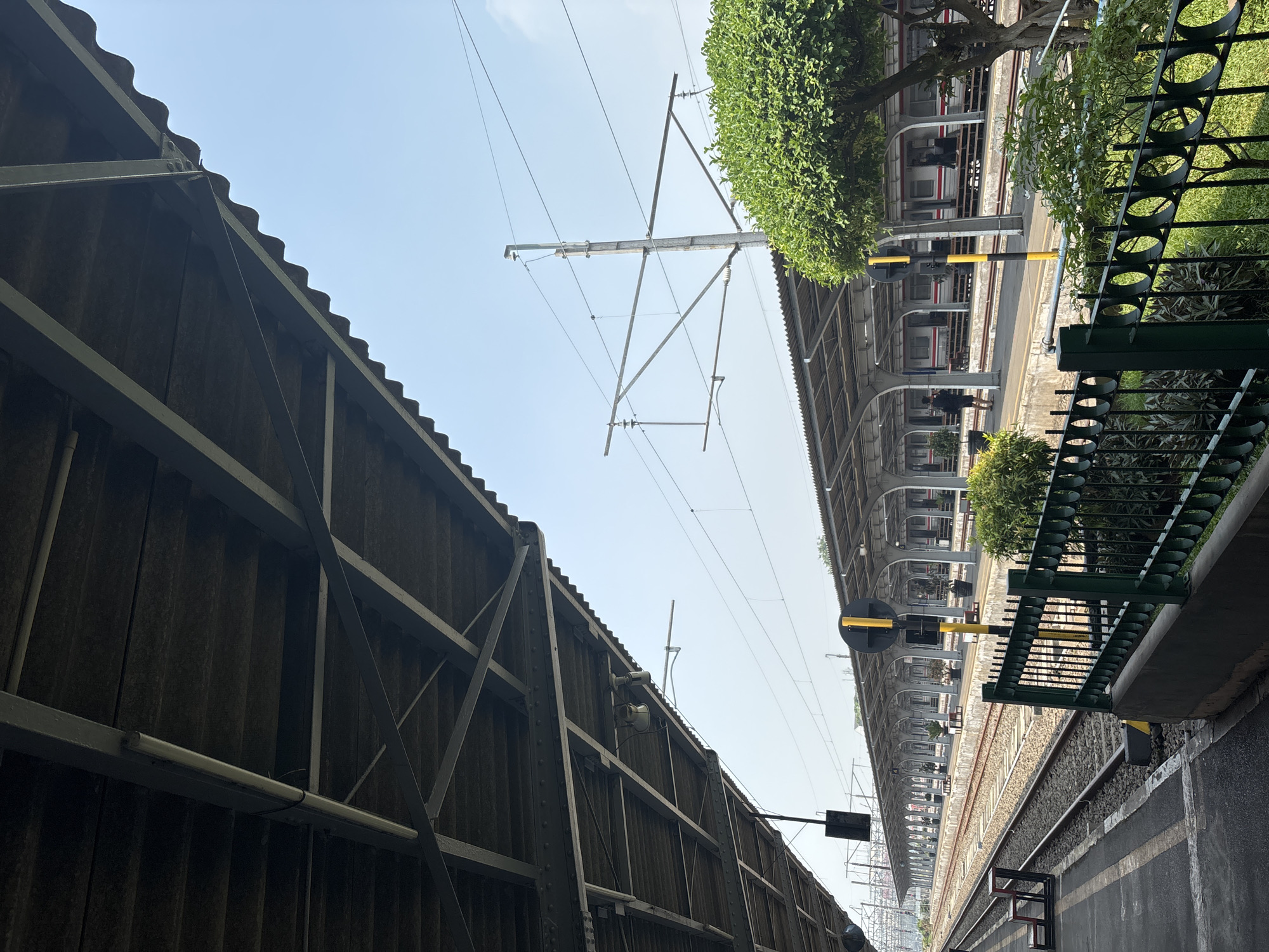 This is a photograph of a train station in West Jakarta, Indonesia, on a clear day. The image is taken from the perspective of a platform, looking down parallel train tracks. A large, corrugated metal roof supported by beams extends over the platform, casting shadows. In the distance, covered platforms with arched supports line the tracks, with trains visible beneath them. The tracks themselves are laid on gravel. A tall, yellow and black striped pole, likely a signal, stands prominently in the foreground. A green metal fence with circular cutouts separates the platform from a grassy area and lush green trees. The sky is a pale blue, indicating fair weather.