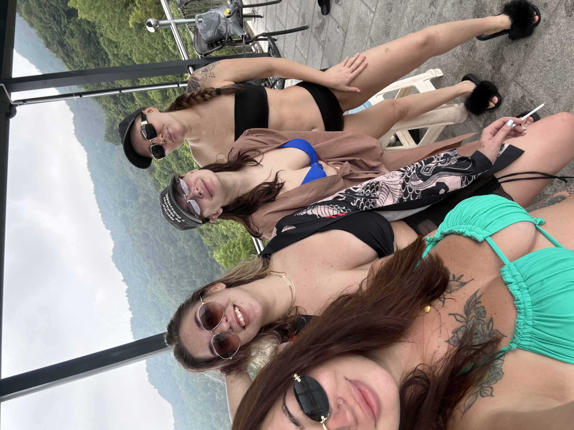 Four young women are posing for a selfie outdoors. They are positioned on what appears to be a balcony or elevated platform overlooking a mountainous landscape covered in lush green trees. The sky is overcast.
The woman on the far left is closest to the camera, her head and upper body filling the lower left portion of the frame. She has long, dark brown hair and is wearing a bright turquoise bikini with ruffled edges and a cutout design. A tattoo is visible on her chest. She is wearing small, oval sunglasses with gold frames and her mouth is slightly open as if she is speaking or smiling. She holds a lit cigarette in her right hand.
Next to her, and slightly behind, is a woman with blonde hair pulled back. She is wearing a black bikini top and large, circular sunglasses with gold frames. She is smiling and has her left hand resting on her head. A thin, gold chain necklace is visible around her neck.
The third woman from the left is wearing a blue bikini top and a light brown, open robe with a patterned design on the sleeves and front. She has dark brown hair and is wearing a black baseball cap with white text. She is looking towards the camera with a slight smile and is wearing thin-framed sunglasses.
The woman on the far right is wearing a black strapless bikini top and black bikini bottoms. She has blonde hair braided into a plait and is wearing large, rectangular sunglasses with a gold frame. She has a tattoo on her upper arm and is looking towards the camera with a neutral expression. She is standing and her left hand is resting on her hip.
In the background, a metal railing and some outdoor chairs are visible. The ground is paved with grey stone tiles. The overall impression is a casual gathering of friends enjoying a scenic view. The mention of Yiwu, China is not visually supported by the image content.