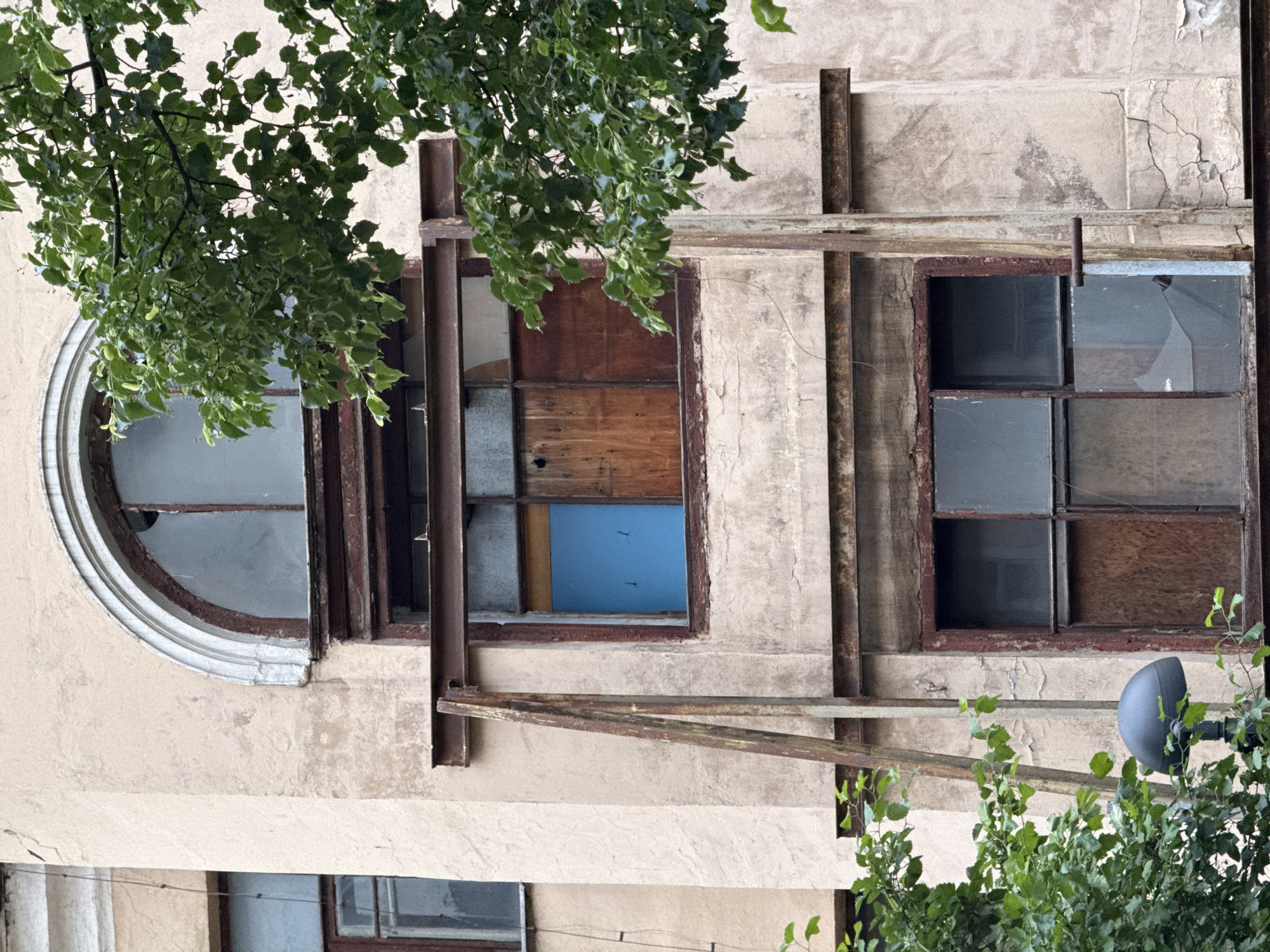 The image shows a building facade with peeling beige plaster. Three windows are visible: a rounded window on the left, a boarded-up window in the middle, and a multi-pane window on the right with broken panes. Rusting metal bars are affixed to the building's exterior. Vegetation partially obscures the view. The location is in Moscow, Russia.