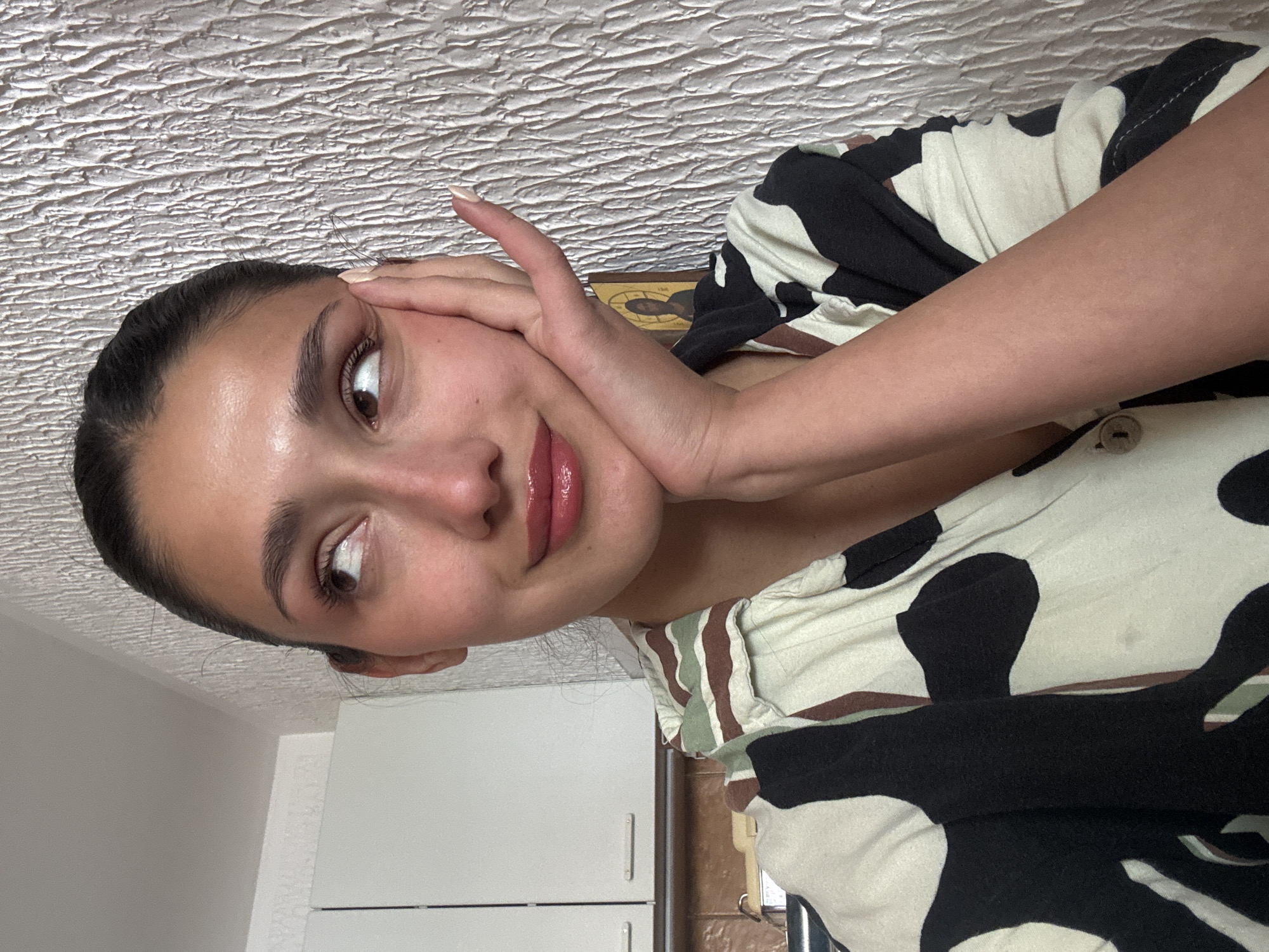 A close-up portrait, rotated 90 degrees counter-clockwise, features a young person (likely female) with dark hair pulled back, looking to their left (viewer's right) with a subtle, reflective expression. Their left hand, with manicured nails, gently touches their cheek. The person has brown eyes, well-defined eyebrows, and glossy pinkish-nude lips. They are wearing a short-sleeved top with an abstract pattern in off-white, black, and hints of brown and olive green.
The setting appears to be indoors, likely a home in Sutomore, Montenegro, during the daytime, suggested by the bright, diffused lighting. The background includes a white, textured popcorn ceiling above their head (when oriented correctly). Behind their right shoulder is a light-colored wall with white kitchen-style cabinets, and behind their left shoulder, a darker wall with what appears to be a small, framed religious icon or picture resting on a surface. No visible text is present in the image.
