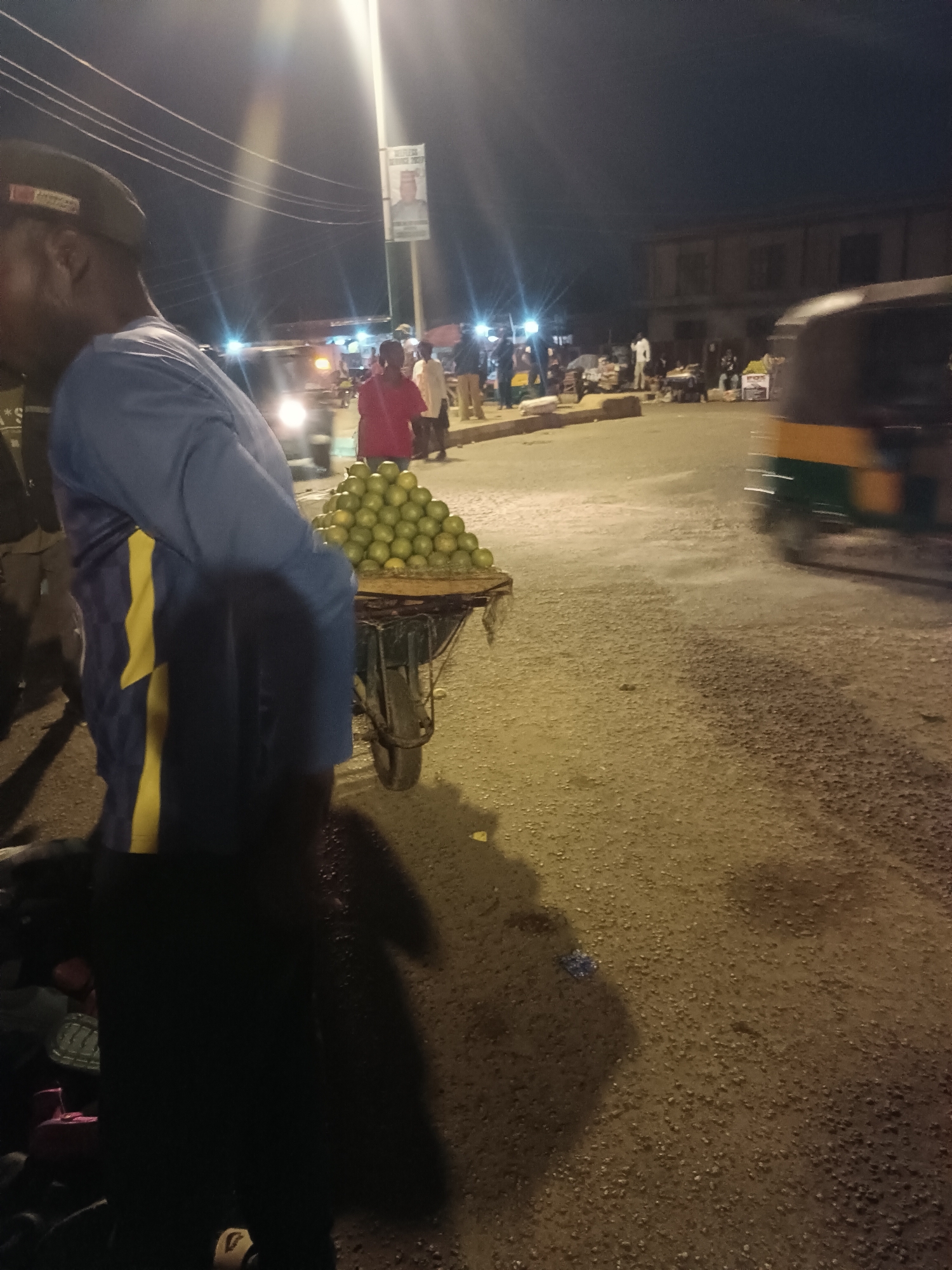 A nighttime scene in what appears to be Bukuru, Nigeria. A man wearing a cap, blue shirt with yellow stripes, and dark pants stands on the left, partially obscuring a cart piled high with green citrus fruit. The cart is parked on a street that seems to be made of gravel. In the background, illuminated by streetlights, are several people gathered. A tuk-tuk drives past on the right side of the road. A political poster is mounted on a light pole in the background. A two-story building is also visible in the background.