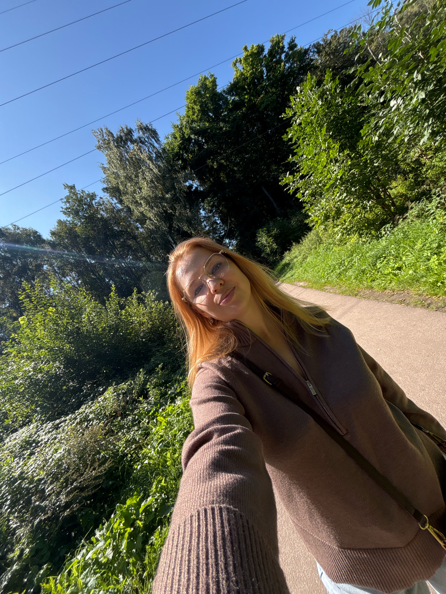 A young woman with blonde hair and glasses is taking a selfie outdoors. She is wearing a brown sweater and appears to be walking on a paved path through a wooded area. The sun is shining brightly, casting shadows and highlighting the greenery. The location is identified as Rusanovo, Russia, with power lines visible in the clear blue sky, suggesting an outdoor setting within or near a populated area.