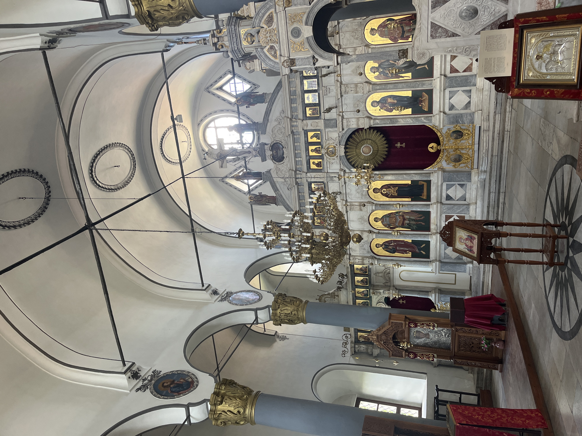 The image displays the ornate interior of an Orthodox Christian church, located in Plovdiv, Bulgaria. The central focus is a grand, multi-tiered iconostasis, intricately carved from light-colored stone or marble with extensive gold leaf accents. It is adorned with numerous icons depicting saints and biblical figures, most featuring golden backgrounds. The Royal Doors, flanked by primary icons, are visible in the lower part of the iconostasis, with a rich red curtain visible behind them.
A large, multi-branched brass chandelier hangs prominently from the high, vaulted ceiling of the nave. The ceiling itself is painted in light tones and features decorative moldings, round and oval medallions with painted religious figures, and dark structural support rods. Natural light illuminates the space through several arched and circular windows located high on the walls.
In the foreground and to the right, two substantial grey columns with elaborate golden Corinthian capitals stand. The floor is tiled with a patterned design, including a large radial motif. Scattered throughout the nave are carved wooden church furnishings, including several lecterns (analoi). One such lectern holds an open book with visible text (unreadable) and a framed icon, possibly depicting St. George and the Dragon, rendered in silver-colored metal against a gold background. Another richly carved wooden stand closer to the iconostasis holds a large, silver-clad icon, likely of the Theotokos. Red fabric drapes over some of the wooden furniture. The church appears empty, and the daylight suggests a time between services.
