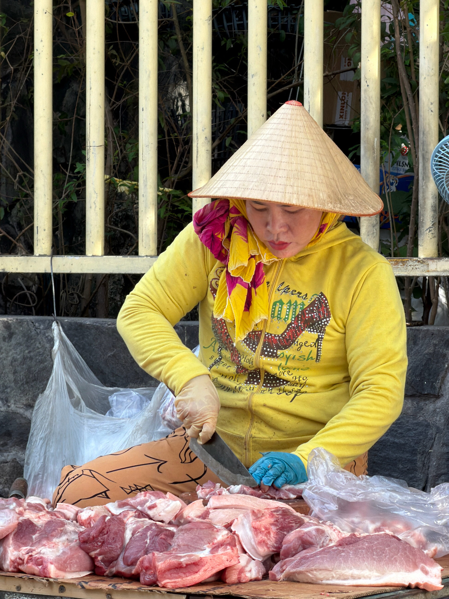 An outdoor scene in Nha Trang, Vietnam shows a vendor, wearing a conical hat, yellow hooded sweatshirt, and light brown pants, preparing raw meat. The vendor is sitting behind a table or stall laden with numerous cuts of pork, some in plastic bags. A large knife is being used by the vendor to cut the meat. The background consists of a yellow metal fence with vertical bars, behind which vegetation is visible. Part of a blue electric fan is also visible on the right side of the frame. The ground in front of the vendor is a textured grey surface. The overall lighting suggests it is daytime.