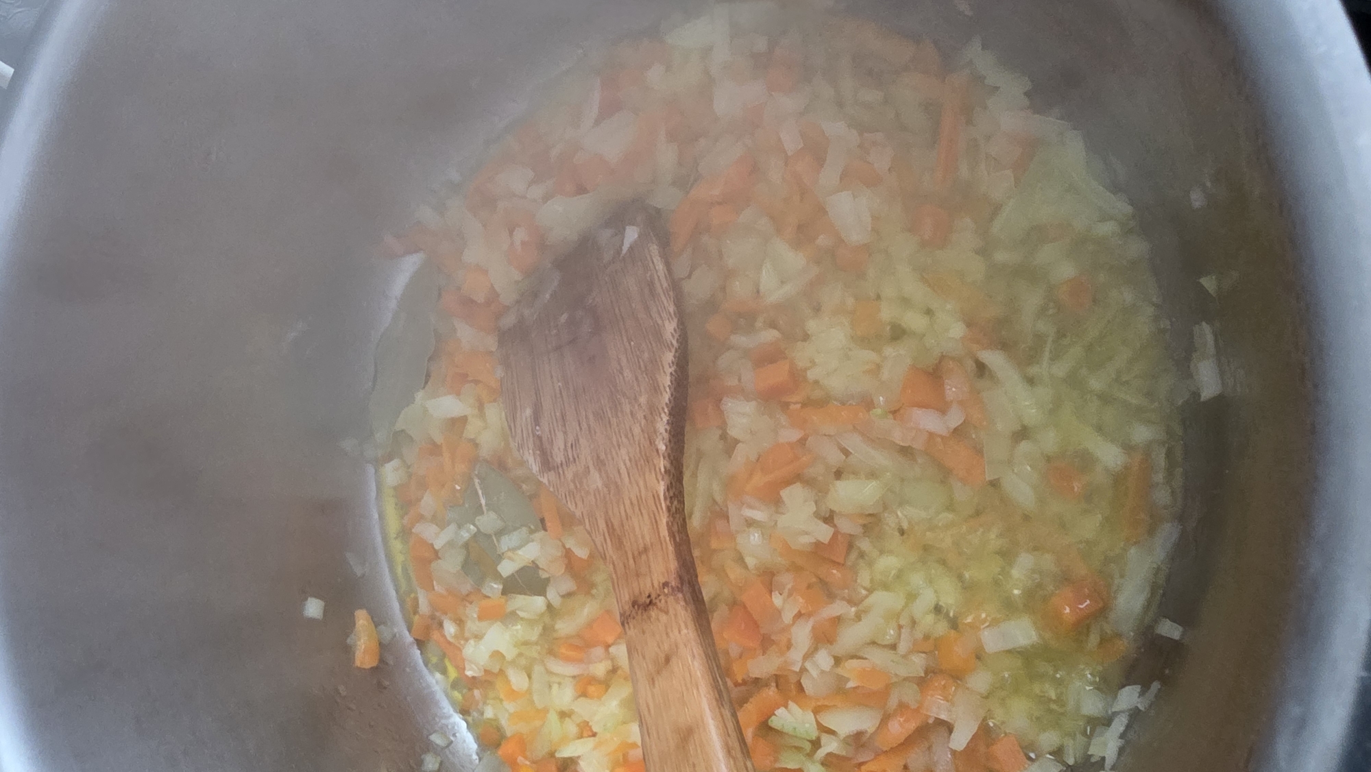 A top-down, medium shot shows a stainless steel pot containing diced onions and carrots being stirred with a wooden spoon. The vegetables are translucent, and some are partially submerged in a yellow liquid, likely cooking oil or stock. Steam is visible rising from the pot's contents. The pot appears to be on a stovetop. The specific location in Blagoevgrad, Bulgaria cannot be determined from this image.