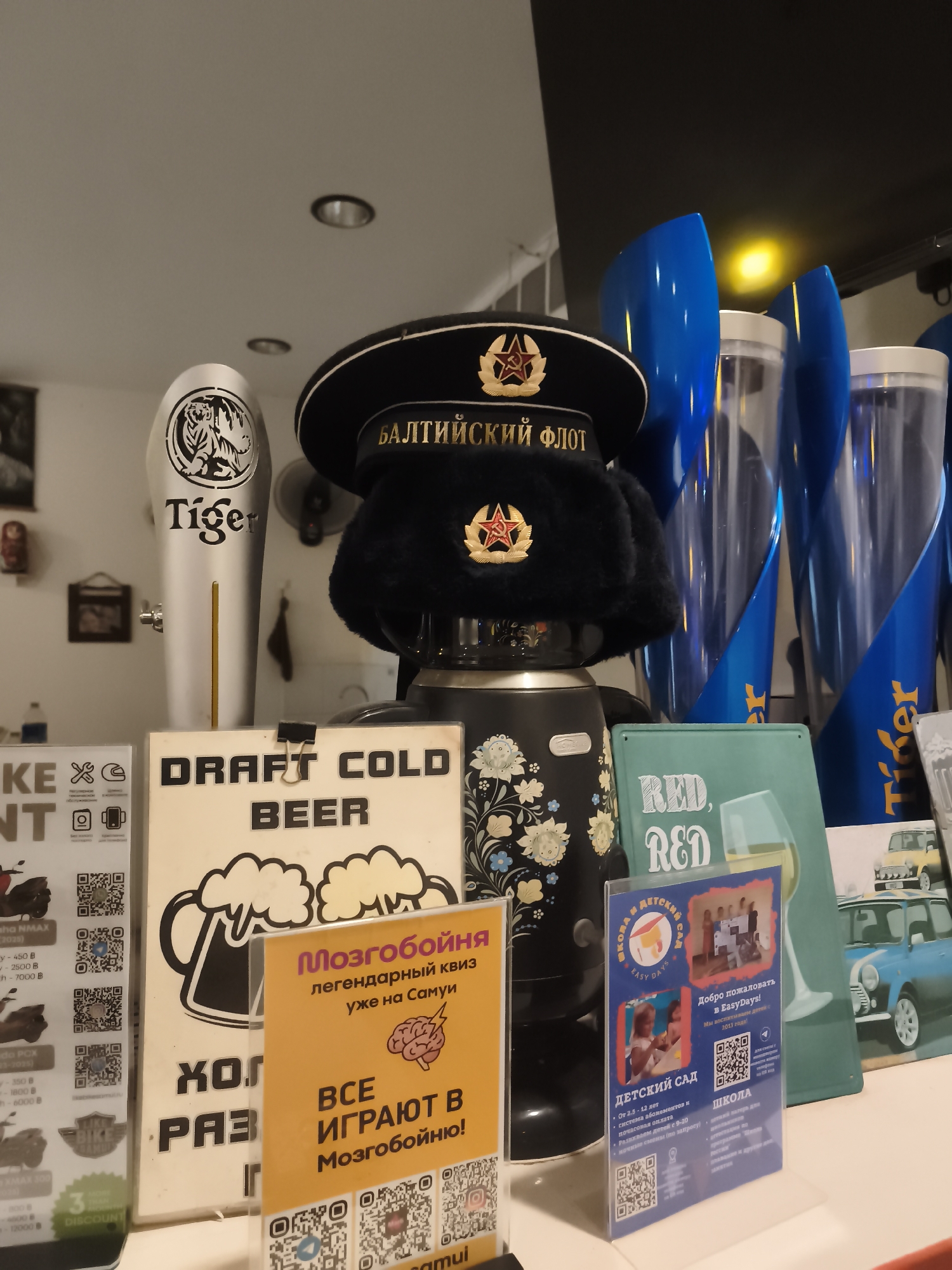 This image appears to be taken at a bar or restaurant in Baan Mae Nam, Thailand. The focus is on a collection of items displayed on a counter.
In the center, a black Soviet-style naval hat with a red star emblem is perched on top of a black cylindrical object decorated with white floral patterns. To the left of the hat is a silver beer tap with a stylized tiger graphic and the "Tiger" logo. Behind the hat and tap are tall, translucent blue containers, possibly for beverages, with "Tiger" branding visible on some.
In front of these items, several signs are displayed. One sign, in black and white, reads "DRAFT COLD BEER" and features an illustration of a frothing beer mug. Another sign, in bright yellow, has Russian text that translates to "Brain Buster - the legendary quiz is already on Samui" and "Everyone plays in Brain Buster!". Below this, a pink sign with Russian text is visible, mentioning "EasyDays" and featuring photos of children.
To the far left, a portion of a price list or menu is visible, showing prices in Thai Baht (B) and model names like "NMAX" and "PCX," suggesting it might be related to motorcycle rentals or sales. A "3 DISCOUNT" sign is also present.
The overall setting appears to be indoors, likely a bar or tourist spot given the variety of items and the presence of the beer tap and signs promoting a quiz and children's activities. The lighting suggests it could be daytime or evening. There are no people prominently visible in the frame. The presence of Russian text on multiple signs suggests a significant Russian-speaking clientele or ownership.