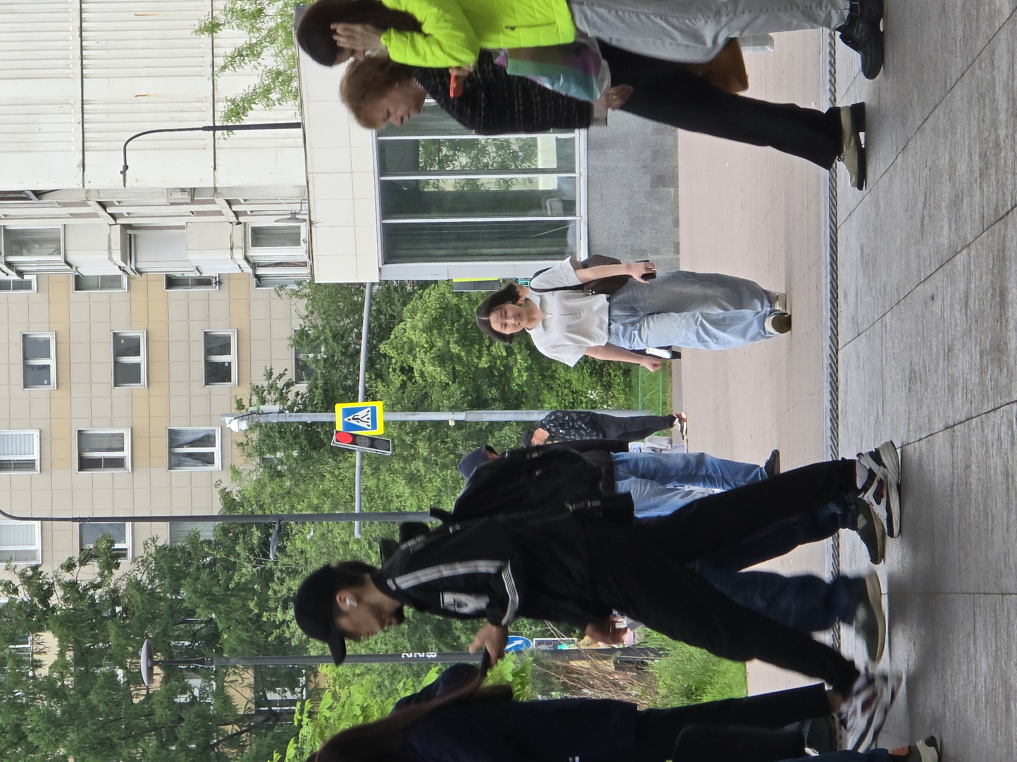 The image depicts a pedestrian street scene in Moscow, Russia. Several people are walking on a sidewalk in various directions. There is a building in the background with multiple windows and a building next to it with larger windows. A crosswalk sign with a red light is visible. The foreground includes individuals of varying ages and attire, suggesting a mix of residents and visitors. Some are focused on their paths, while one person in the center is smiling at the camera. The environment consists of trees and greenery, indicating a city park or garden.