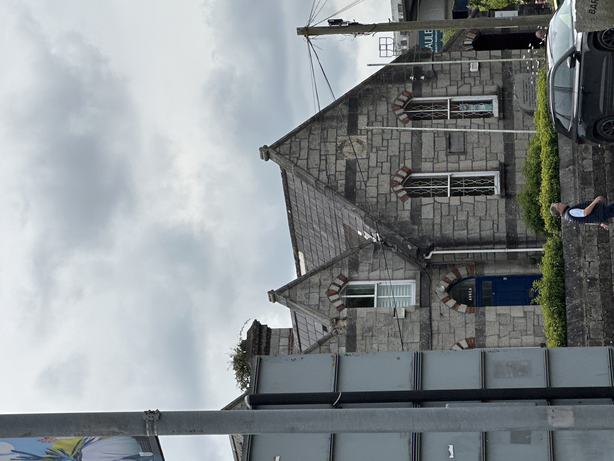 The image captures a cloudy day scene in Carlow, Ireland, focusing on the stone facade of a building with a distinctive pitched roof. The building features several arched windows with intricate metal grilles and white frames. A bright blue door is visible, with the word "AVOCA" written above it in white lettering. Lush green bushes line the front of the building. To the right, a utility pole with a network of wires stands prominently. In the foreground, a dark-colored car is parked on the street, and a man in a dark vest and white shirt is walking away from the camera towards the left. A large, grey, rectangular structure, possibly a billboard or signage, is partially visible on the left side of the image, obscuring part of the building. The overall impression is one of a historic building situated within an urban environment.