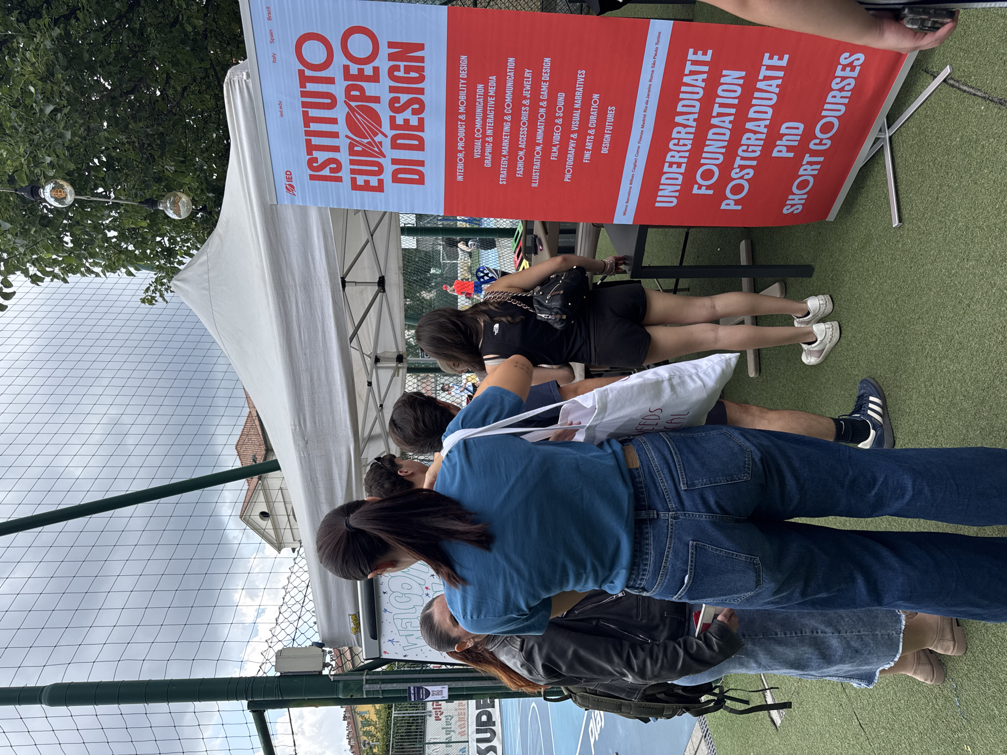This image depicts an outdoor promotional event for the "Istituto Europeo di Design" (IED) in an urban area of Milan, Italy. The scene is set on a patch of green Astroturf, under a partly cloudy sky.
In the foreground and midground, a group of approximately six young adults are gathered around a white canopy tent that houses an informational booth. A prominent red and blue banner for IED stands on the right, listing various design courses such as "Interior, Product & Mobility Design," "Visual Communication," "Fashion, Accessories & Jewelry," and "Illustration, Animation & Game Design." It also specifies program levels: "Undergraduate," "Foundation," "Postgraduate," "PhD," and "Short Courses." The banner includes a list of IED campus locations, with "Milano" being the first listed in Italy, alongside other cities like Rome, Madrid, and Rio de Janeiro.
The central figure is a woman seen from behind, wearing a teal t-shirt, blue flared jeans, and a white tote bag partially obscuring text that reads "NEEDS CAL." Her dark hair is tied in a ponytail. To her left, another woman with dark hair, wearing a black leather jacket and a light denim midi-skirt, is partially visible, carrying a black backpack. To the right of the central figure, a person in a black tank top and black shorts, carrying a black shoulder bag with a chain strap, stands facing the booth. Other individuals are partially visible or obscured by the central figures.
Under the white tent, dark tables are set up, and a "Welcome" sign with colorful doodles is visible on a white surface behind the central woman. Two decorative light bulbs, one clearly featuring a Bitcoin symbol, are suspended by cords from a tree branch above the tent.
The event area is separated by a green chain-link fence from a blue basketball court visible in the background, where a QR code for "BASKET" is affixed. Beyond the fence and court, a mix of urban architecture is visible, including buildings with traditional red tiled roofs and a classical pediment, alongside a modern building with a glass facade.