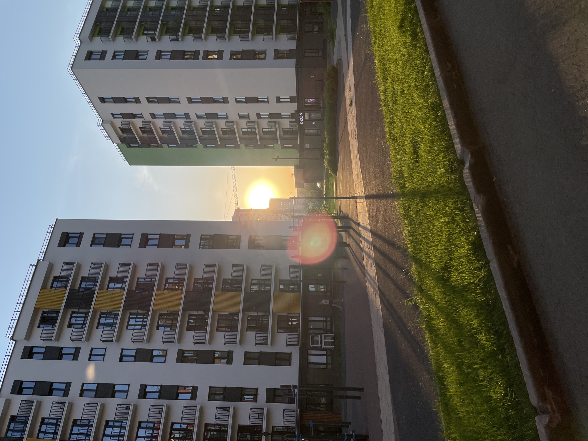 The image shows two modern apartment buildings in Pervomaysky, Russia, at sunset. The buildings are white with dark-framed windows and balconies. One building features green and dark gray accents, while the other has yellow and dark gray. The setting sun is visible between the buildings, causing a lens flare. A paved walkway runs between the buildings, bordered by a strip of green grass. Shadows from streetlights and other objects stretch across the walkway. An "OZON" sign is visible on one of the buildings. A bench and street furniture are visible in the foreground.