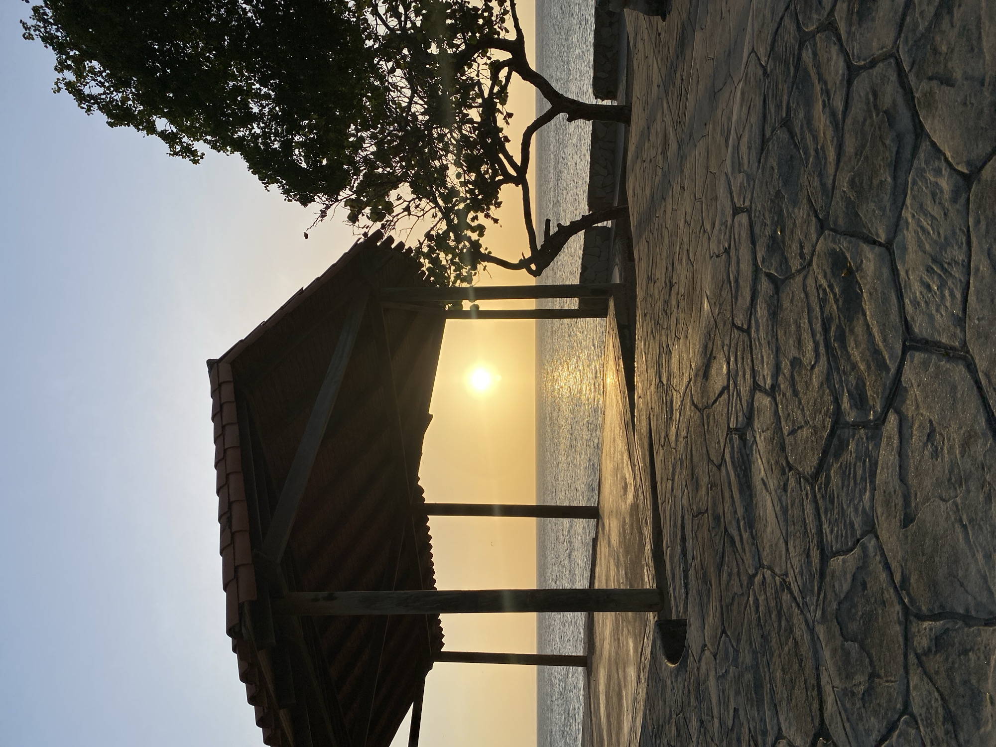 The photograph captures a sunset at Juan Griego, Venezuela. A small, open-sided wooden gazebo with a tiled roof is in the foreground, partially obscuring the view. Beyond the gazebo, the sun is setting over a body of water, reflecting light onto the surface. A tree with sparse foliage stands beside the water. The foreground is a textured stone patio or walkway. No people are visible in the scene.