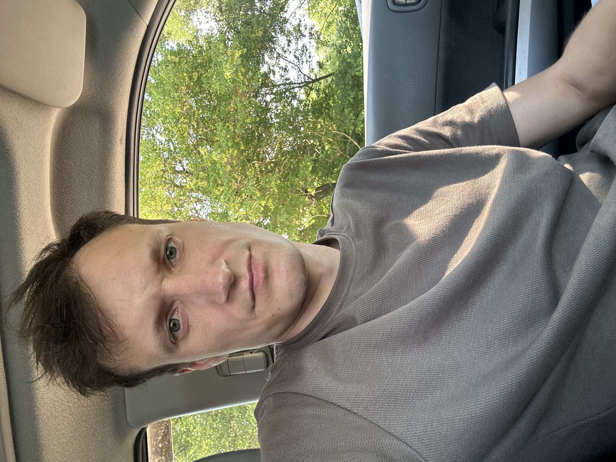 A man is captured in a selfie taken inside a vehicle. He has short, dark, messy hair and light blue eyes. He is wearing a grey t-shirt. The interior of the car is visible, including the headliner and a portion of the car door. Through the car window, lush green trees and foliage are visible, suggesting an outdoor, possibly rural or suburban, setting. The lighting indicates it is daytime, with sunlight casting shadows. The specific location of Makiv, Ukraine, is not discernible from the visual information presented in the image.