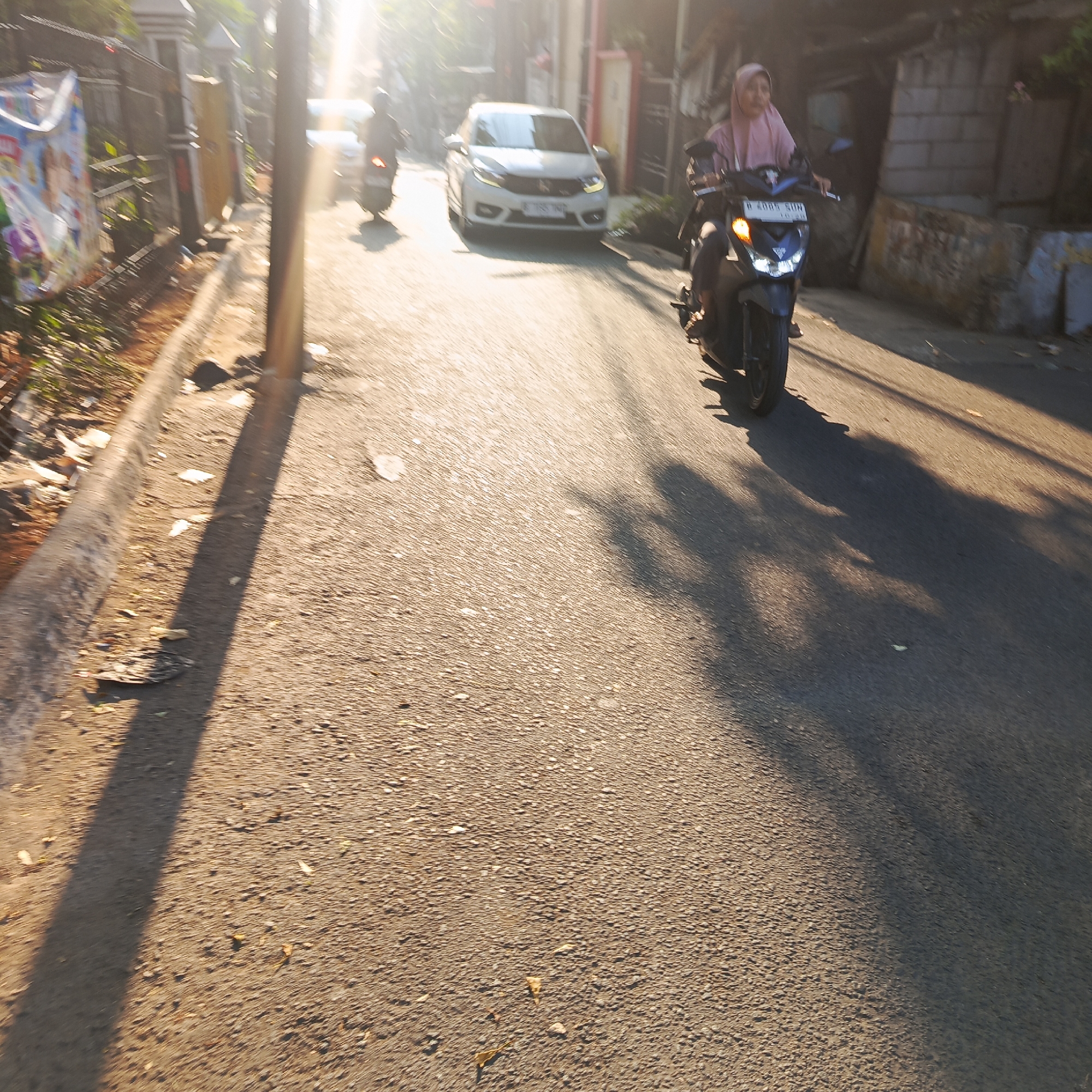 The image captures a sunlit street scene in South Jakarta, Indonesia, possibly during the morning or late afternoon given the long shadows. In the foreground, the asphalt road is textured with small stones and speckled with fallen leaves and debris. A prominent shadow from a pole or structure falls across the road from the left.
In the mid-ground, a white car is visible, moving away from the viewer. To the right of the car, a person is riding a motorcycle, also moving away from the viewer. This motorcyclist is wearing a pink hijab and is partially obscured by the angle of the shot and motion blur. The motorcycle's front light is illuminated, indicating it is in use. Further in the background, another motorcycle is visible, also in motion.
The left side of the image shows the edge of the road with a concrete curb. Behind the curb, there are some plants and a fence. A large, colorful banner, possibly an advertisement or poster, is hanging on the fence, partially unfurled. The buildings in the background appear to be typical of a residential or commercial area in a city, with some showing brickwork and corrugated metal roofing. The overall atmosphere is bright, with the sun creating a lens flare effect and contributing to the motion blur of the moving vehicles.
