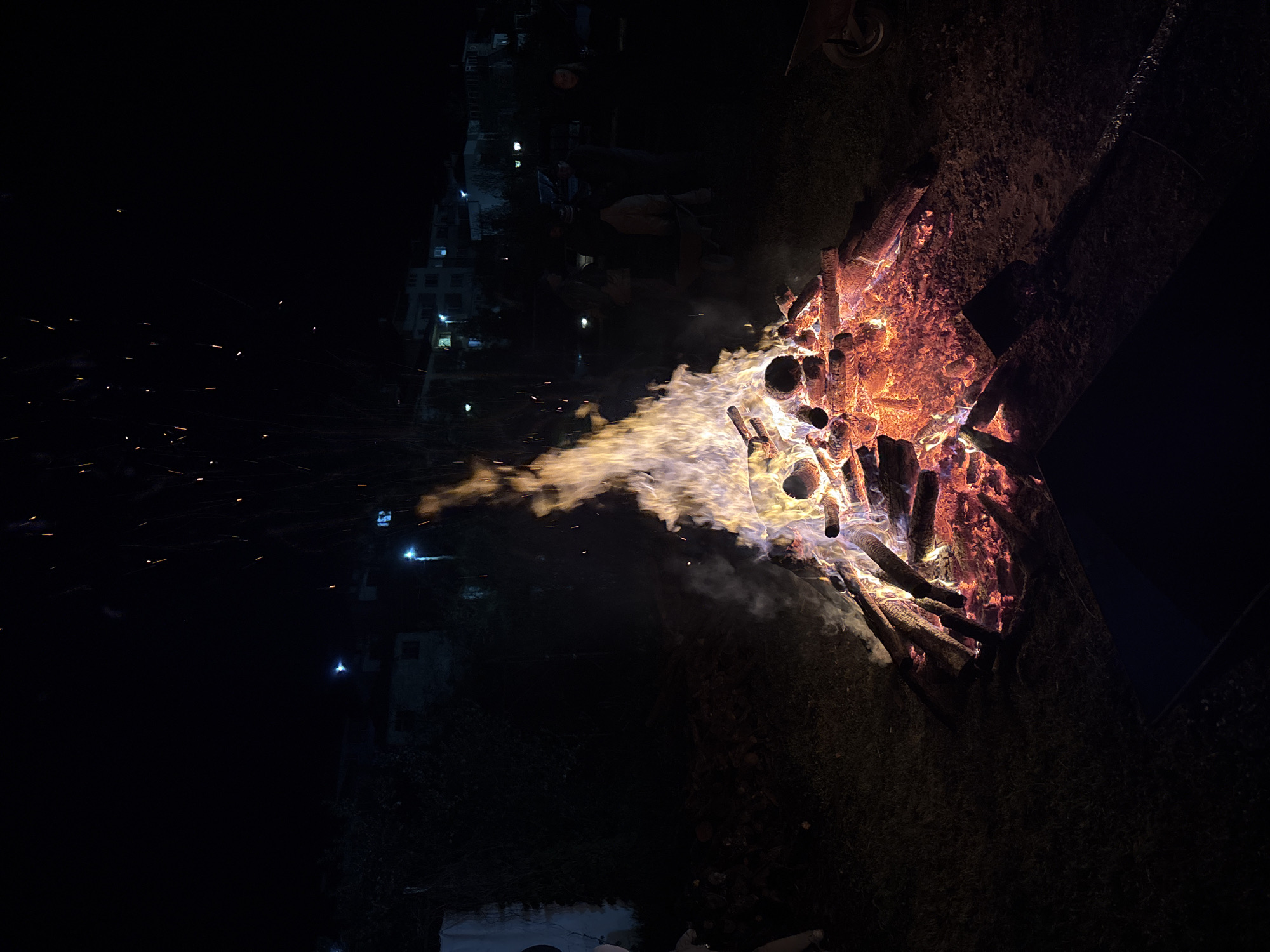 A vibrant bonfire blazes at night, dominating the right side of the frame. Intense orange, red, and yellow flames engulf numerous logs, casting a bright, flickering light. Wisps of smoke rise from the fire, partially illuminated by the intense heat, while a shower of bright, glowing sparks arcs upwards and to the left against the dark sky. The immediate area surrounding the fire appears to be dark earth or grass. In the distant left background, the faint outlines of buildings with scattered illuminated windows suggest a settlement, indicating human presence. These distant lights imply the scene is taking place near a town or village, specifically Zlatograd, Bulgaria, as provided by the context. The time of day is clearly night, with the deep darkness creating a strong contrast with the brilliant fire. No specific weather conditions are discernible, but the smoke and sparks suggest calm air. There are no identifiable people or visible text in the image. The scene conveys a powerful and lively outdoor event, likely a gathering, centered around the warmth and spectacle of the large fire.
