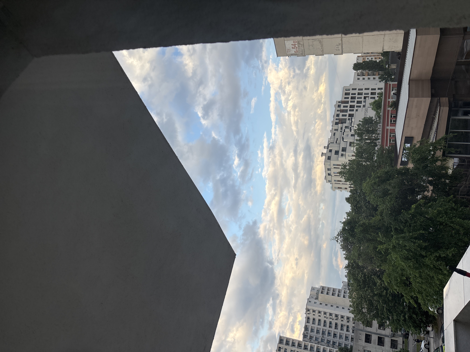 The image presents an urban landscape view, likely captured from a building or balcony, looking outwards into a partially cloudy sky and a cityscape. The foreground is dominated by dark grey, angular concrete structures, possibly a wall or architectural element of the viewpoint, framing the lower-left and top edges of the scene.
Through this architectural frame, the sky is visible, featuring a mix of clear blue patches and layers of white and grey clouds, illuminated by what appears to be late afternoon or early evening sunlight. On the right side of the frame, numerous multi-story buildings populate the midground and background. These structures exhibit varying architectural styles, with some appearing older and others more modern, predominantly in light white or off-white tones with visible windows. Lush, green trees are interspersed among the buildings, adding natural elements to the urban setting.
No people are visible in the scene. The time of day is suggested by the soft, directional light on the clouds and buildings. Visible text on one of the buildings, "Б.54В" (B.54V in Cyrillic), along with partially visible "I Y S M" (likely part of "ЛУЙСМ" or a similar word in Cyrillic), points towards a location in an Eastern European country, consistent with the provided context of Sofia, Bulgaria. The image captures a static, everyday urban vista under a dynamic sky.