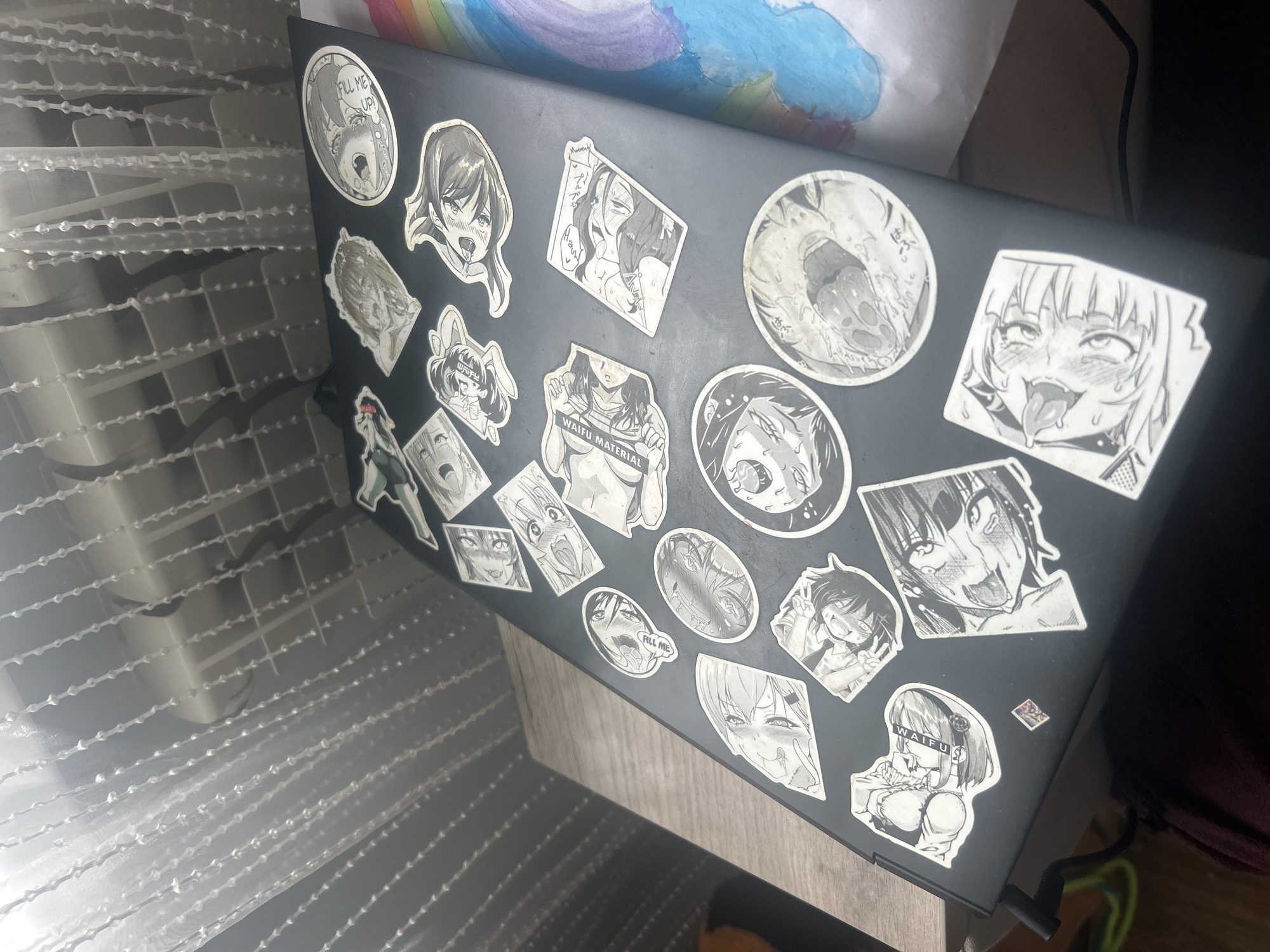 The image shows a black laptop with various anime-style stickers applied to its surface. The stickers depict characters in a black and white, manga-like art style. The laptop is open and placed on a wooden surface. In the background, a white curtain with vertical beaded strands hangs over what appears to be a window or radiator. To the right of the laptop, a child's drawing of a rainbow is partially visible. The lighting suggests an indoor environment, possibly a room. There is no discernible information in the image to indicate a specific location within the city of Супонево, Russia.