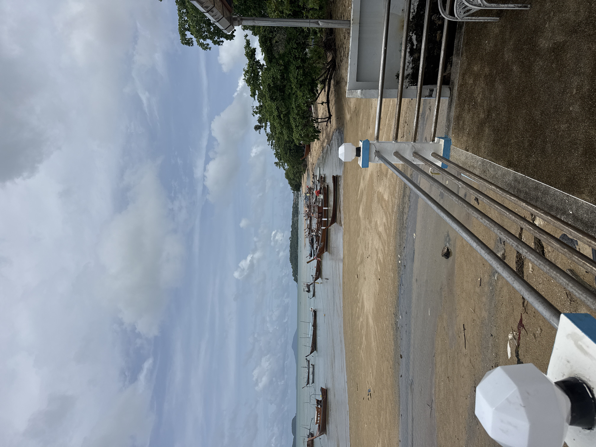 An overcast day frames a tropical beach scene in Rawai, Thailand. A sandy shore slopes gently into the calm, shallow sea, where numerous wooden longtail boats are moored. Some of the boats are partially submerged, their colorful hulls resting on the wet sand. In the distance, hazy islands with tree-covered slopes dot the horizon. The foreground features a concrete walkway with a metal railing, a white hexagonal lamp post, and a small building with a white overhang. Lush green trees line the edge of the beach behind the boats, creating a natural border. The sand is marked with some debris, including pieces of plastic and seaweed, and puddles of water reflect the cloudy sky.