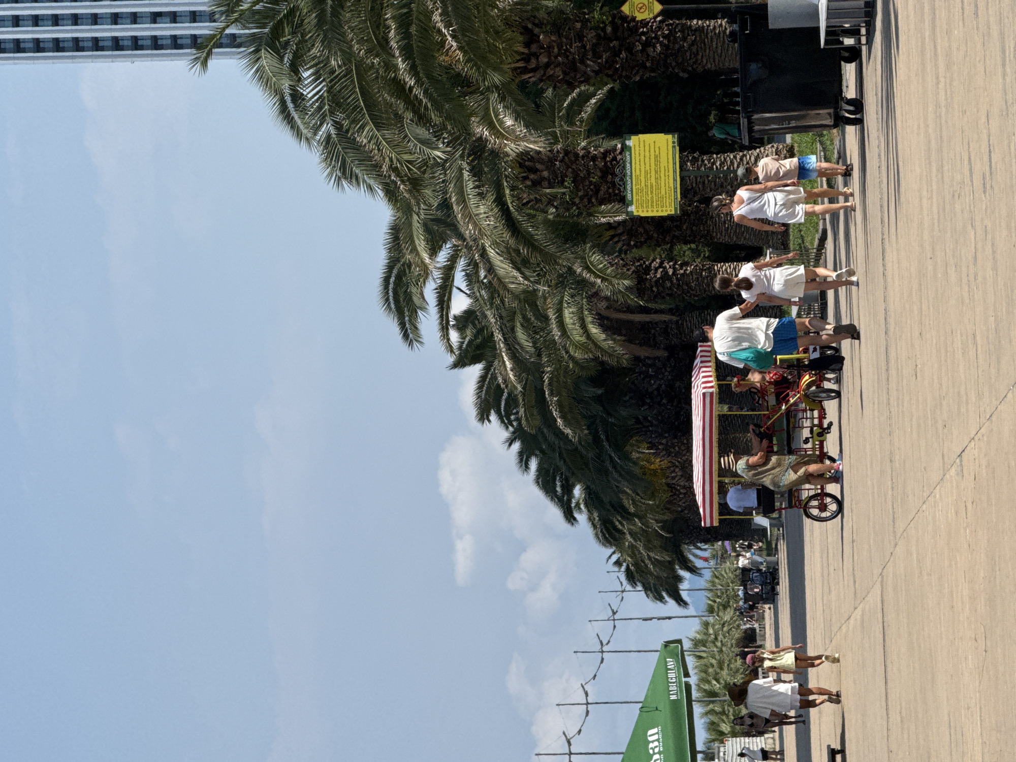 In an outdoor scene in Unknown, Unknown, a group of people walk along a paved walkway lined with palm trees. The sky is a pale blue with scattered clouds. On the right side of the image, a tall, modern building with many windows is visible behind the palm trees. In the foreground, a family of four walks away from the camera. The father wears a white t-shirt and blue shorts, the mother wears a white tank top and white shorts, and their two children, a boy and a girl, wear light-colored clothing. A yellow sign with text in a foreign language is mounted on a pole near the palm trees. To the right of the sign is a large black trash bin with wheels and a metal bench. In the mid-ground, a multi-person pedal vehicle with a striped canopy is stopped, and several people are around it. To the left of the pedal vehicle, another family walks on the walkway, with a woman wearing a hat and white clothing and a young girl in a yellow top and shorts. A green awning with text is visible further to the left, and a tall building with many windows rises in the background on the far right. The overall impression is of a sunny day at a tourist destination.