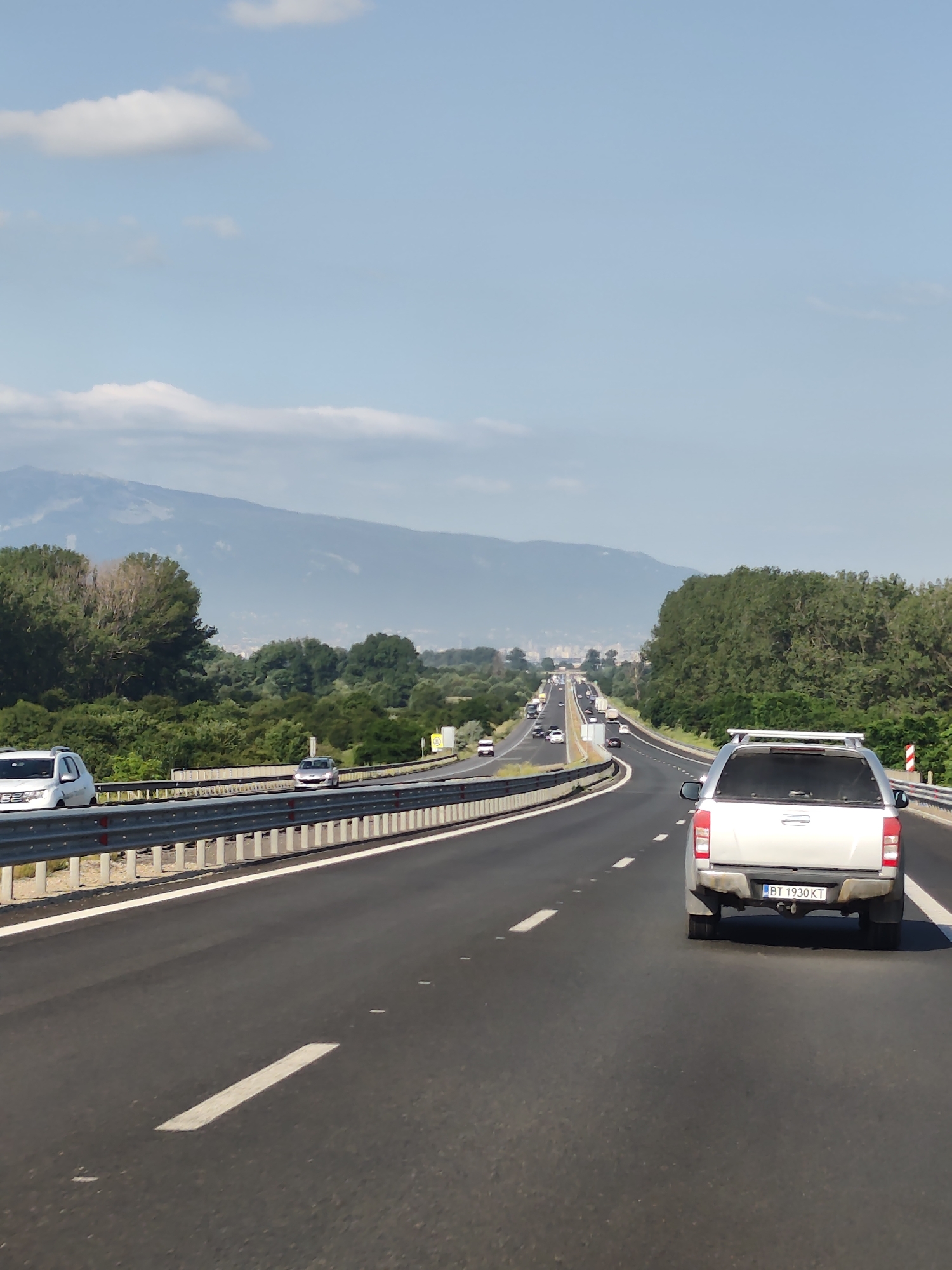 The image shows a highway with traffic on a sunny day. The highway has multiple lanes and is bordered by trees on both sides. In the foreground, a silver pickup truck with the license plate "BT 1930 KT" is visible, driving in the right lane. Other vehicles are present on the highway, with some in the distance. In the background, mountains are visible under a clear blue sky with scattered clouds. The location is identified as the city of Dolni Bogrov, Bulgaria.