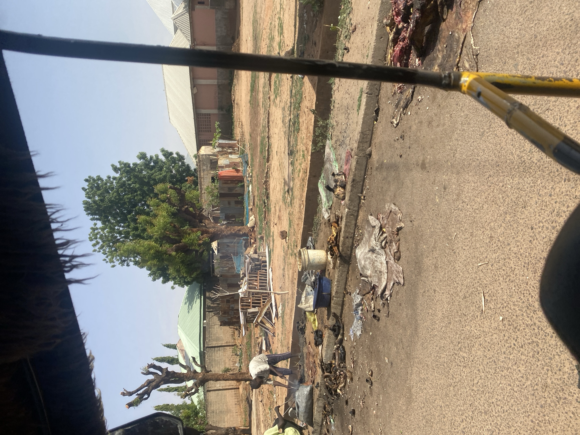 The image shows a street scene in Gombe, Nigeria, likely taken from inside a moving vehicle. Two men are working by the roadside, appearing to be processing animal hides. Debris and litter are scattered along the roadside and there is a stack of wooden crates near the men. Other buildings are visible in the background.